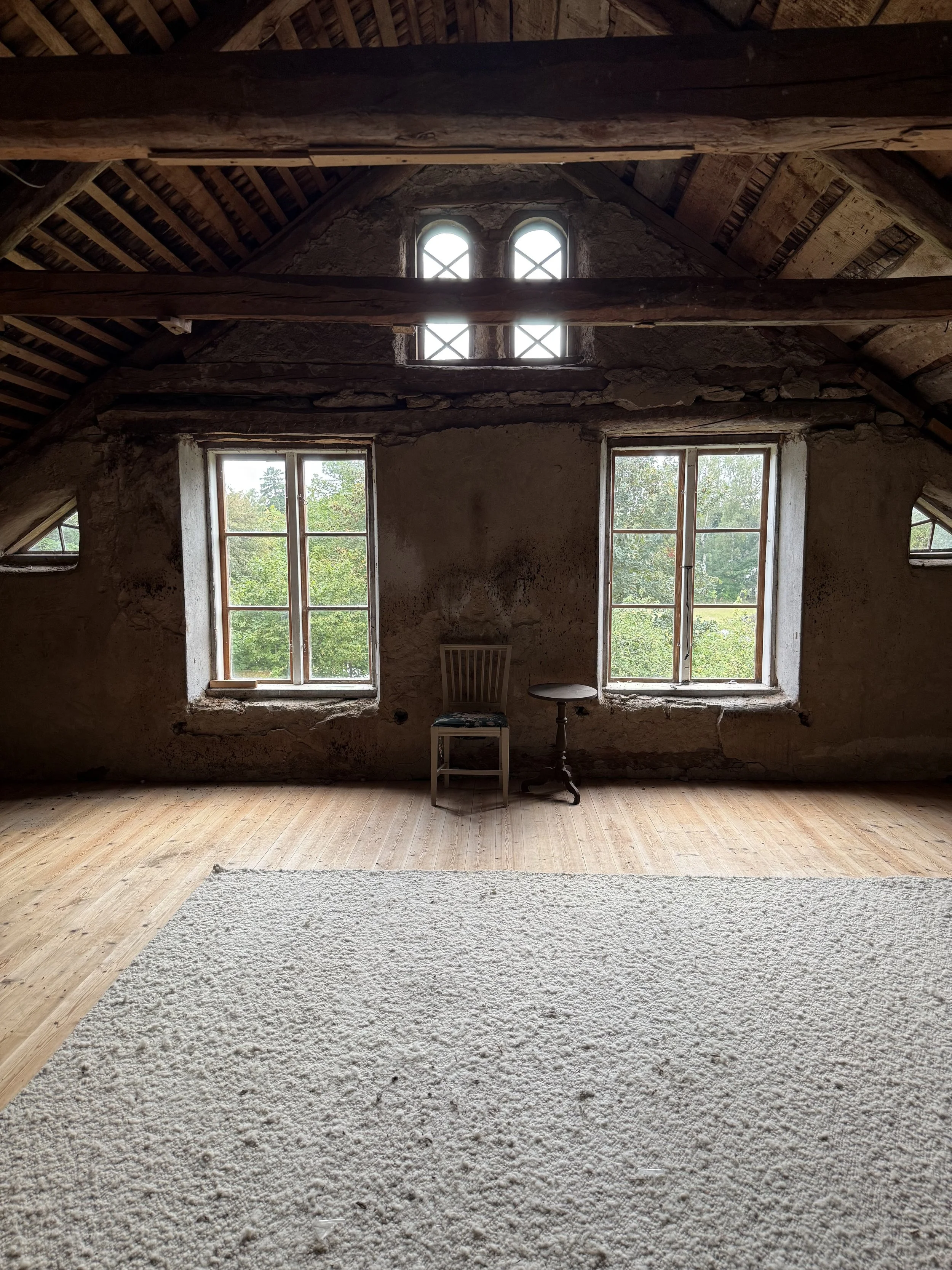 Intérieur rustique d’une ancienne maison en pierre, avec poutres apparentes, fenêtres en bois, chaise et petite table sur un plancher ancien.