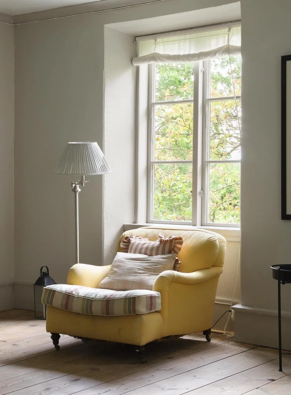Fauteuil jaune avec repose-pieds rayé près d’une fenêtre, accompagné d’un lampadaire dans un intérieur lumineux.