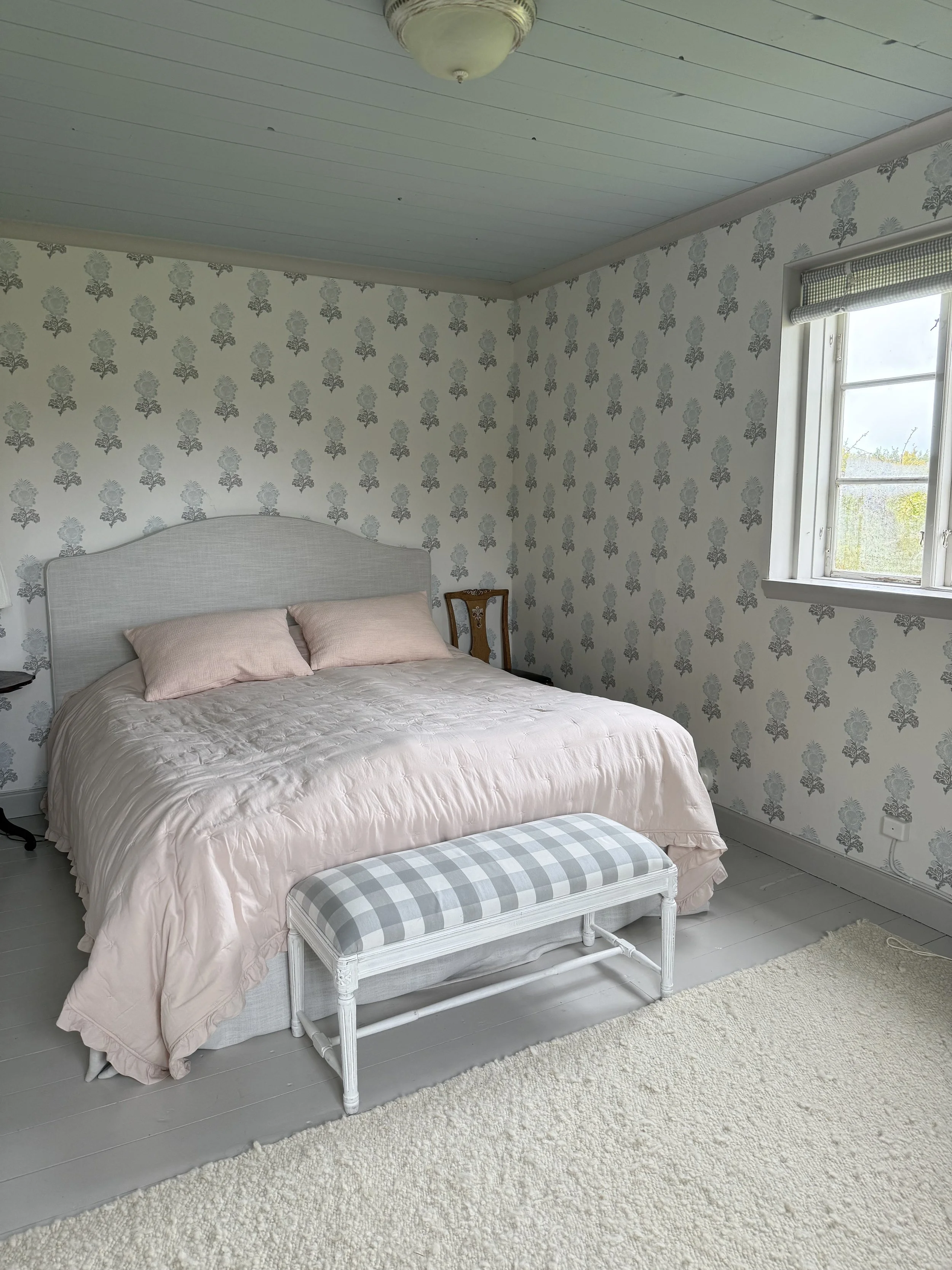 Chambre lumineuse avec lit double rose, banc capitonné à carreaux, papier peint floral, fenêtre et tapis blanc.