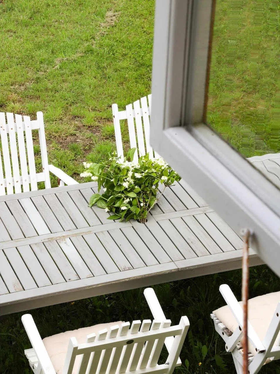 Terrasse en bois avec chaises Adirondack blanches et plante fleurie, vue depuis une fenêtre.