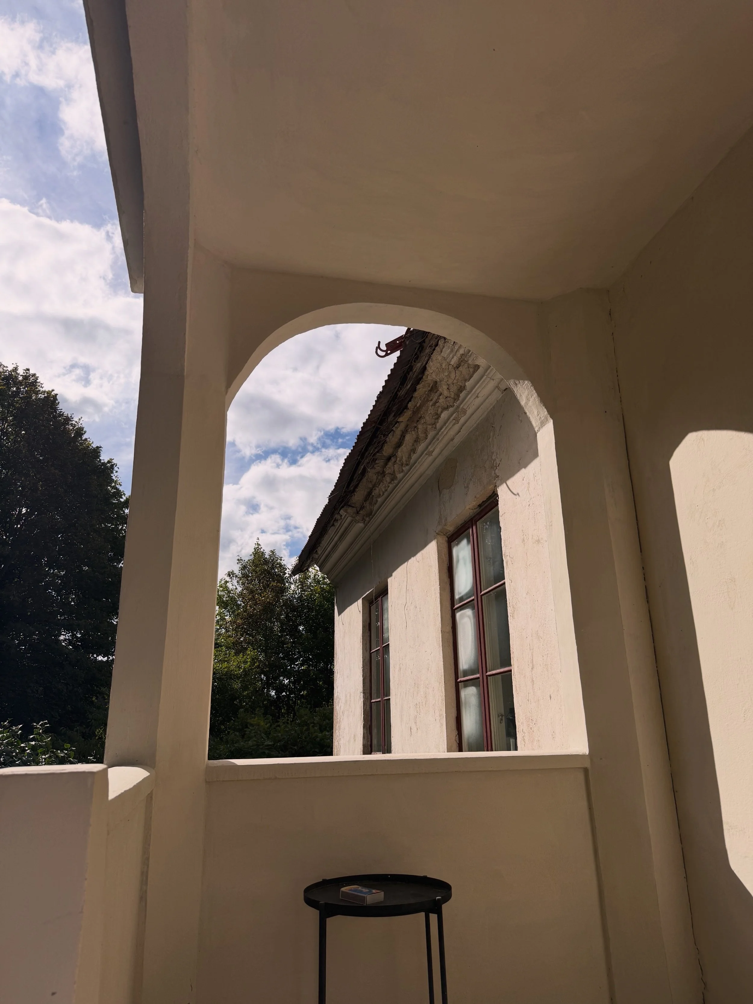 Porche aux arches blanches devant une ancienne maison, avec une petite table noire et un paquet posé dessus.