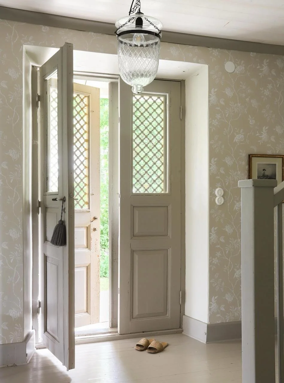 Entrée de maison avec portes ouvertes à treillis décoratif, suspension en verre et pantoufles au sol.