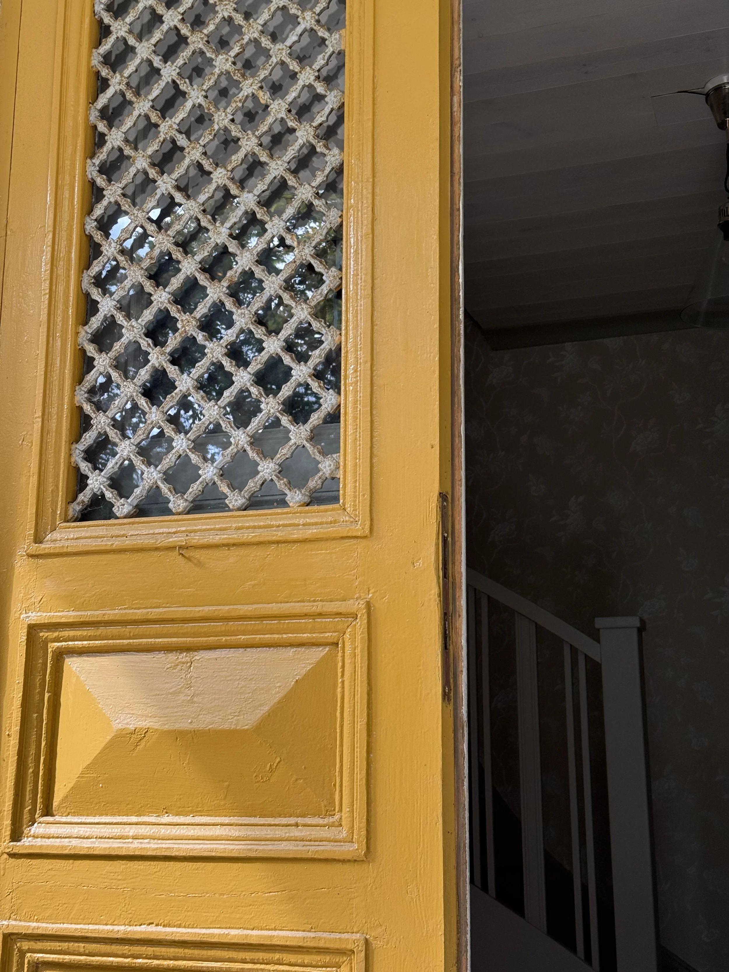 Porte jaune avec fenêtre grillagée en treillis métallique rouillé, donnant sur un escalier sombre.