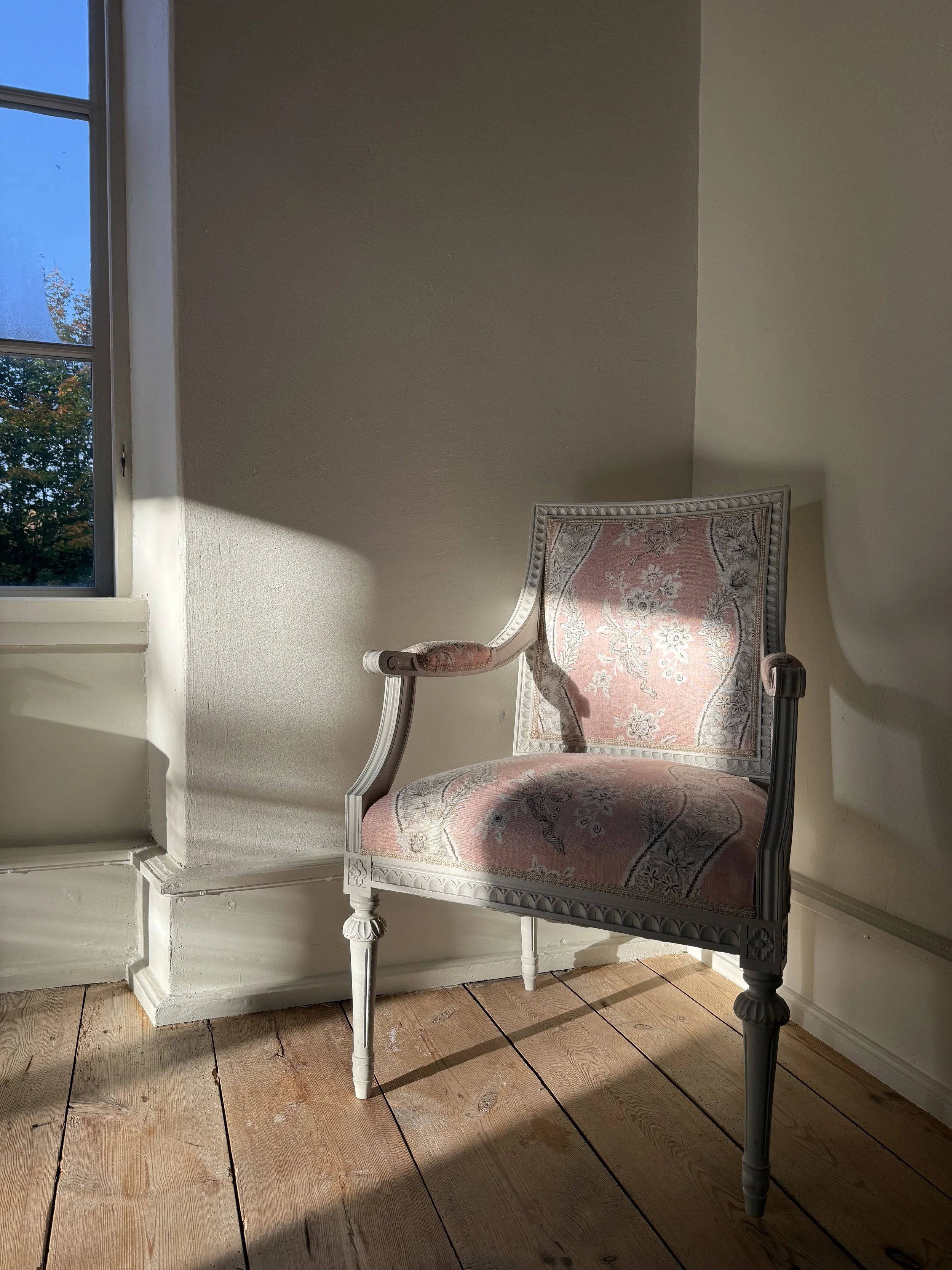Fauteuil ancien en bois sculpté, recouvert d’un tissu rose à motifs floraux, près d’une fenêtre dans un intérieur baigné de soleil.