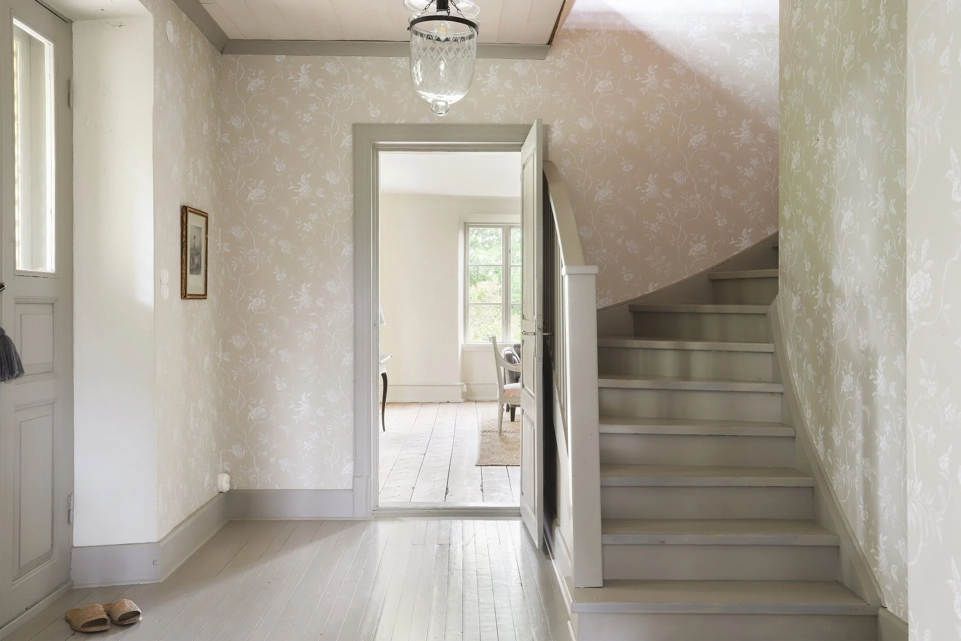 Entrée lumineuse avec papier peint floral, escalier peint en blanc, parquet clair et porte ouverte vers une pièce voisine.