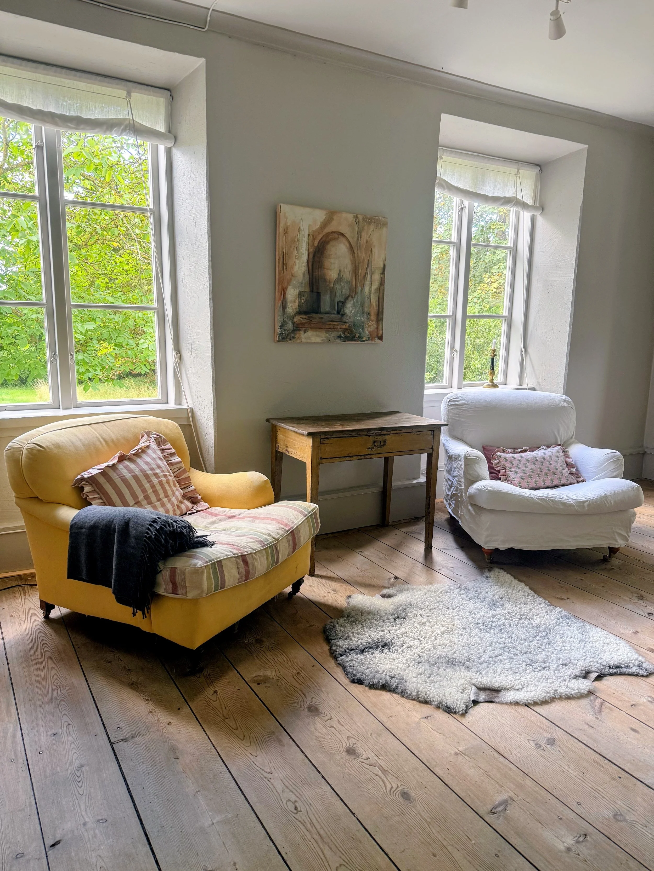 Salon lumineux avec fauteuil jaune, fauteuil blanc, table en bois, tableau abstrait, fenêtres donnant sur la verdure et tapis blanc sur parquet.