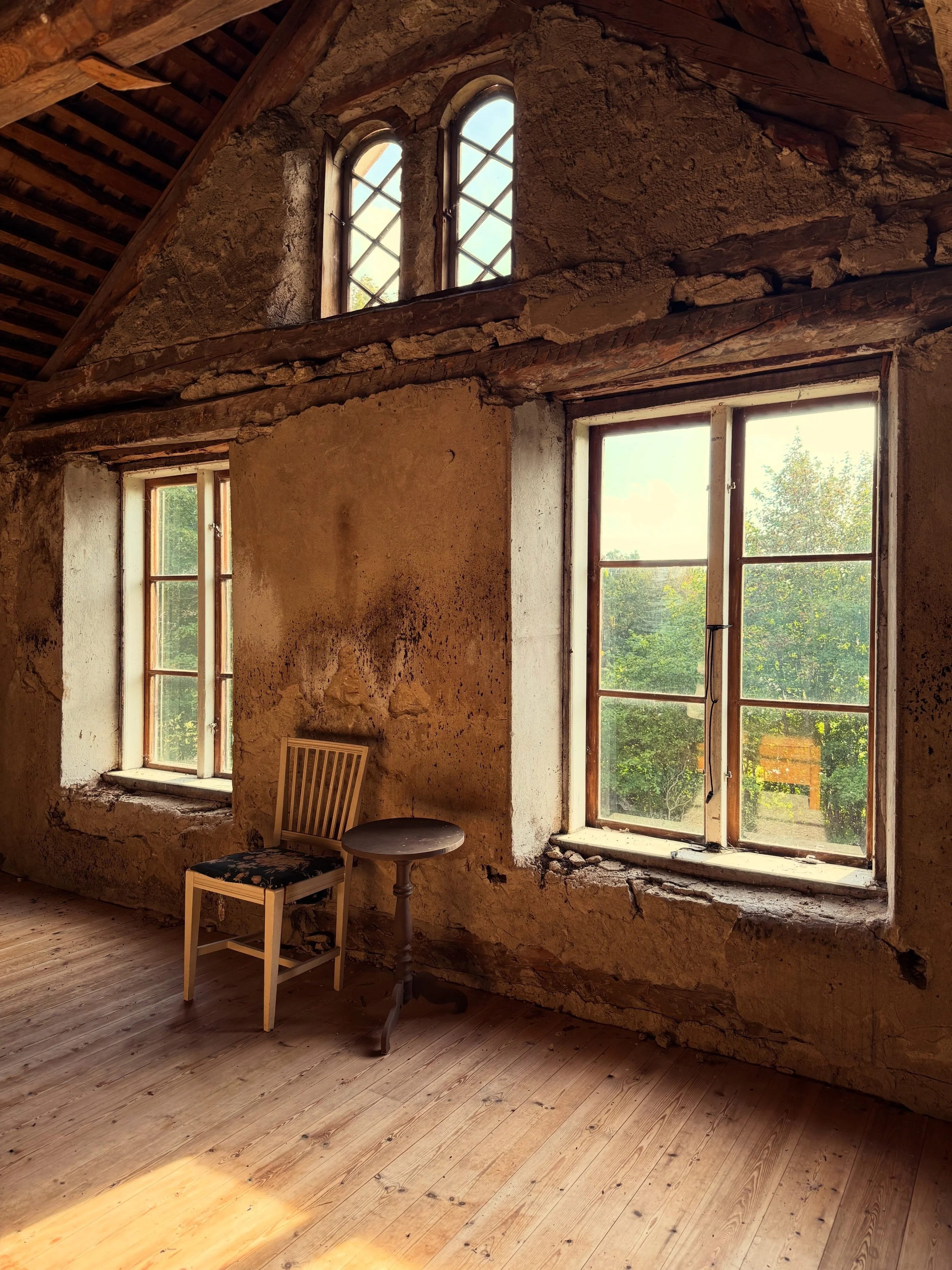 Intérieur rustique d’une maison ancienne aux murs en torchis, poutres apparentes, fenêtres en bois et parquet, avec une chaise et une petite table.