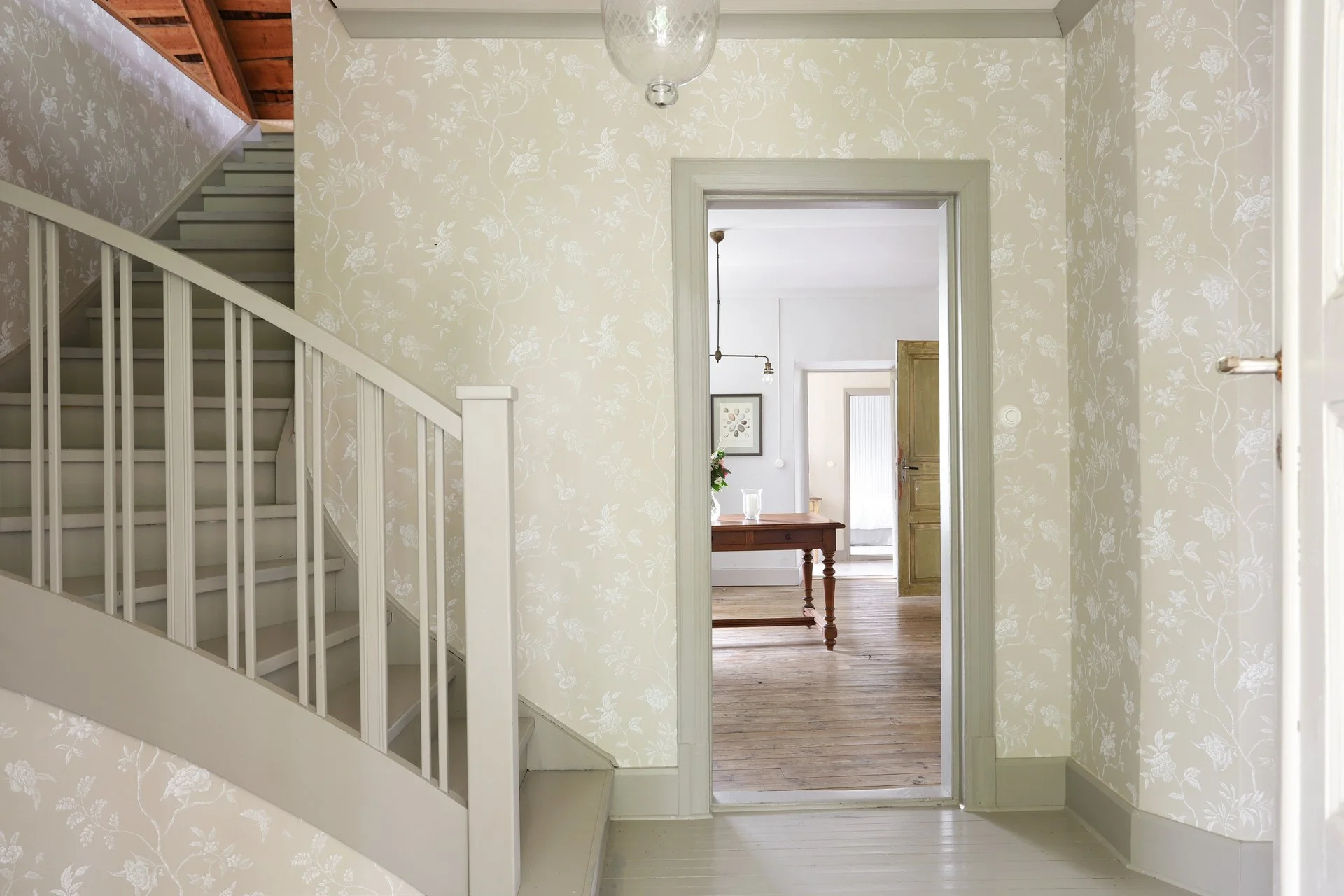 Entrée d’une maison avec escalier peint en blanc, papier peint floral et porte ouverte donnant sur une pièce au parquet en bois.