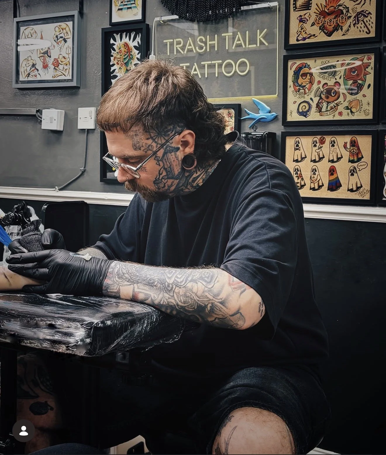 A tattoo artist with tattoos and earrings working on a tattoo in a tattoo studio. The background features framed tattoo art and a neon sign that reads 'TRASH TALK TATTOO.' The artist is focused on his work, wearing black gloves.