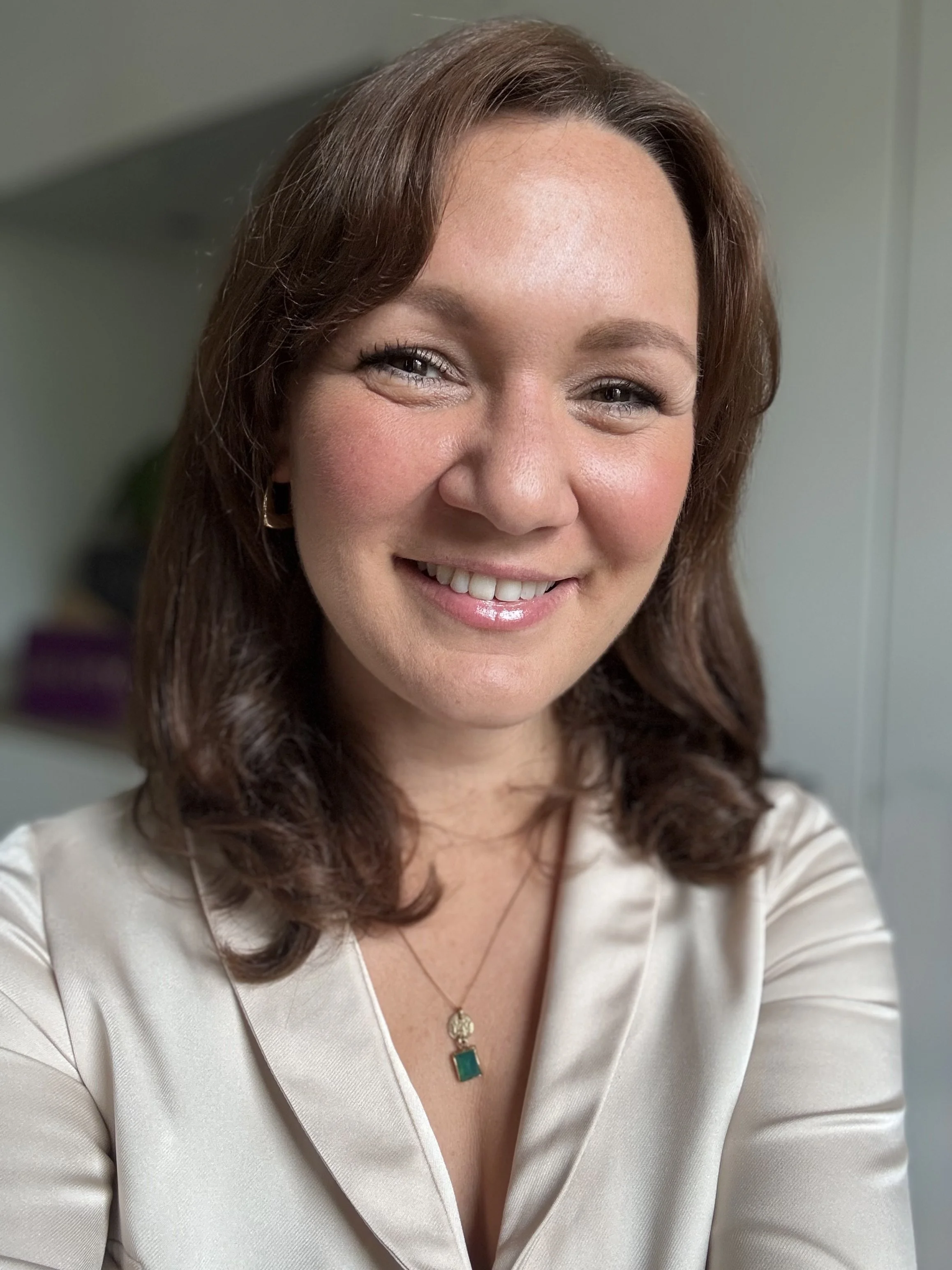 A woman with brown, wavy hair smiling at the camera, wearing a white satin blouse and a gold necklace with a green pendant.
