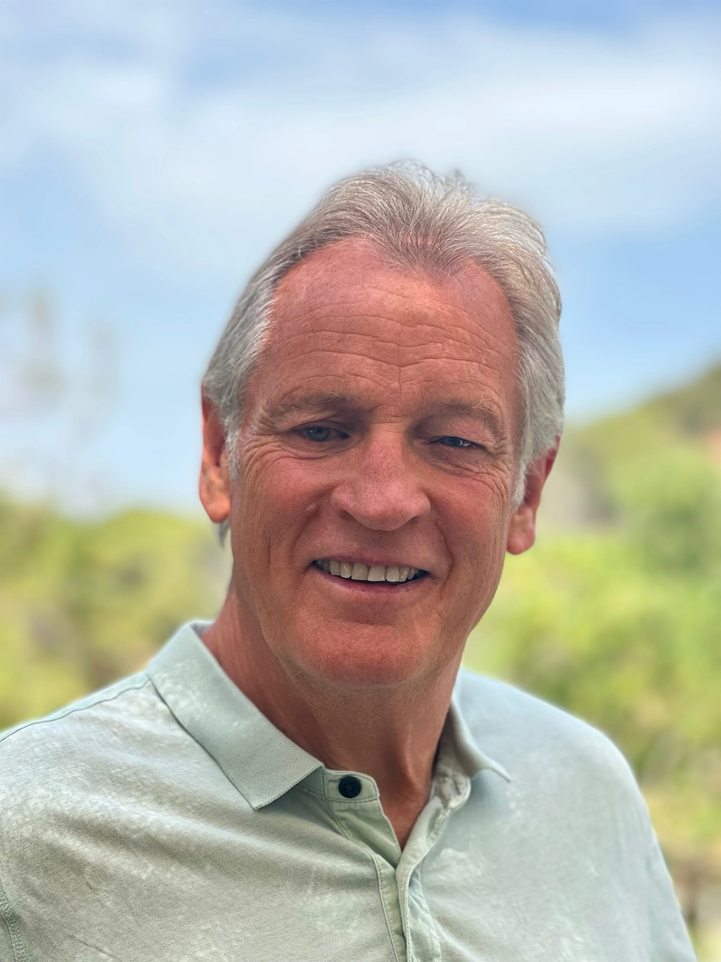 Close-up photo of an older man smiling outdoors on a sunny day, wearing a light-colored polo shirt with a background of blurred greenery and sky.