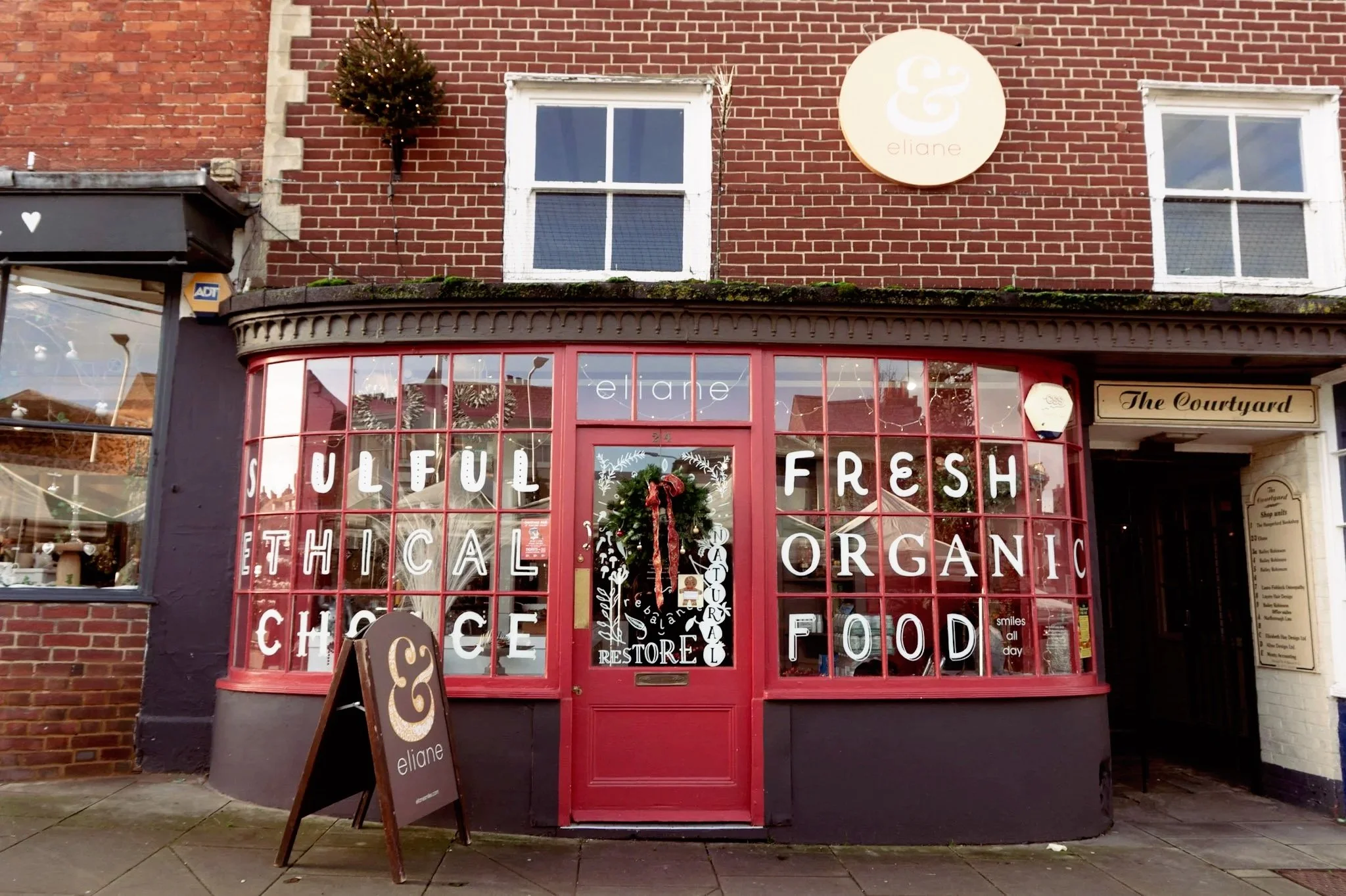 The storefront of a shop named 'eliane' with large red-framed windows displaying signs for organic, ethical, chocolate, and food products. There is a small chalkboard sign outside and a Christmas wreath on the door. The shop has a brick exterior with two upper windows, a round sign with a cloud and the word 'eliane,' and a black-painted section with a small Christmas tree decoration on a bracket.
