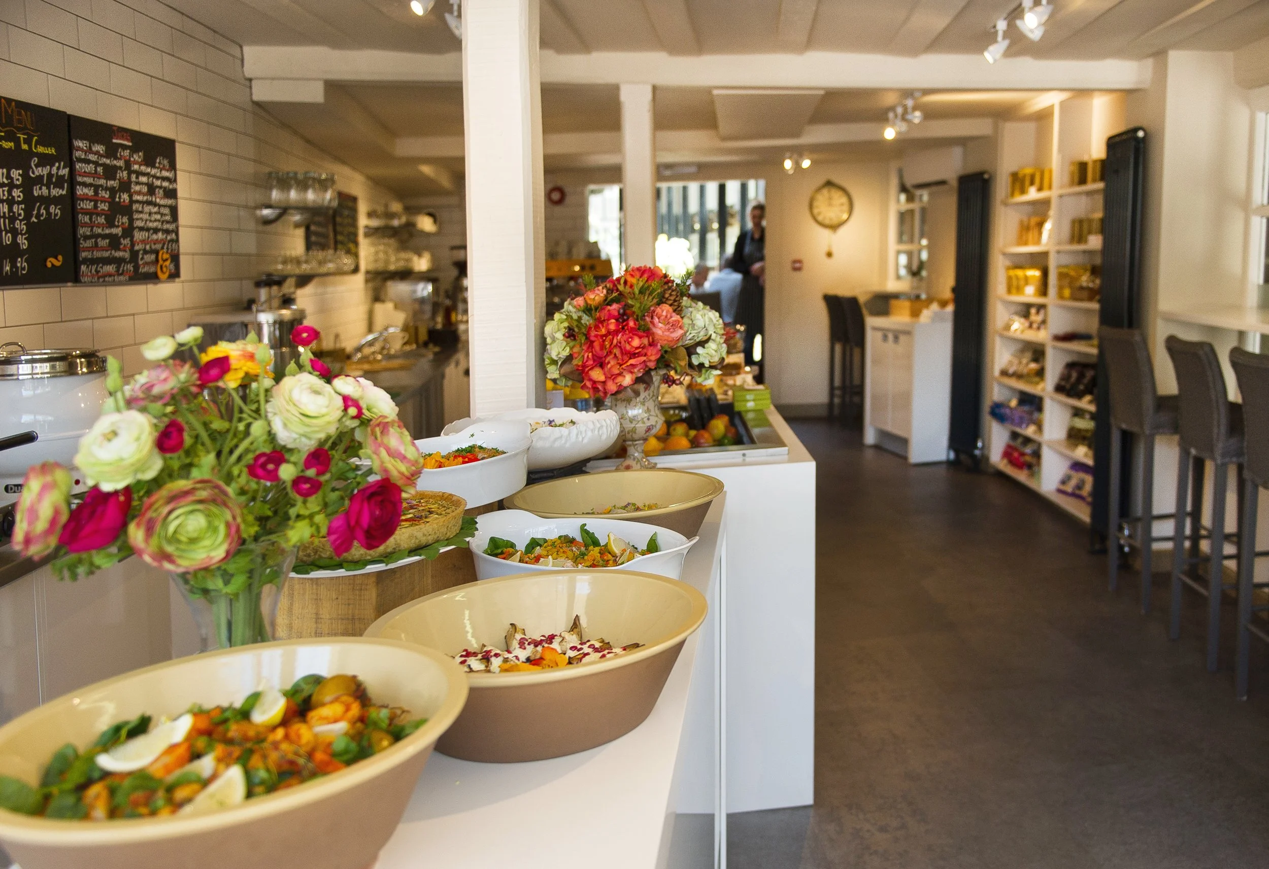 Interior of a cozy cafe or restaurant with a display of salads and floral arrangements in bowls, wooden shelves filled with goods, and a person in the background near a large clock on the wall.