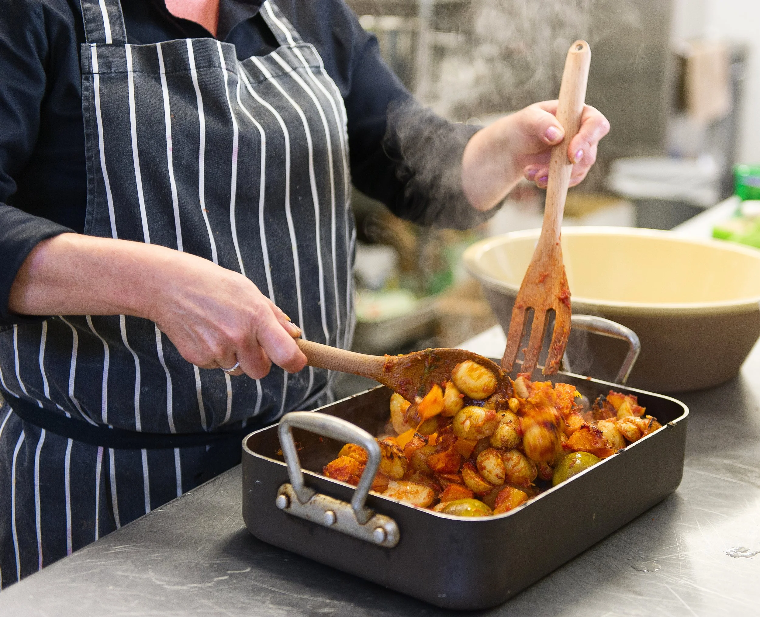 Person wearing a striped apron stirring a dish filled with roasted vegetables, including potatoes, carrots, and Brussels sprouts, in a kitchen.