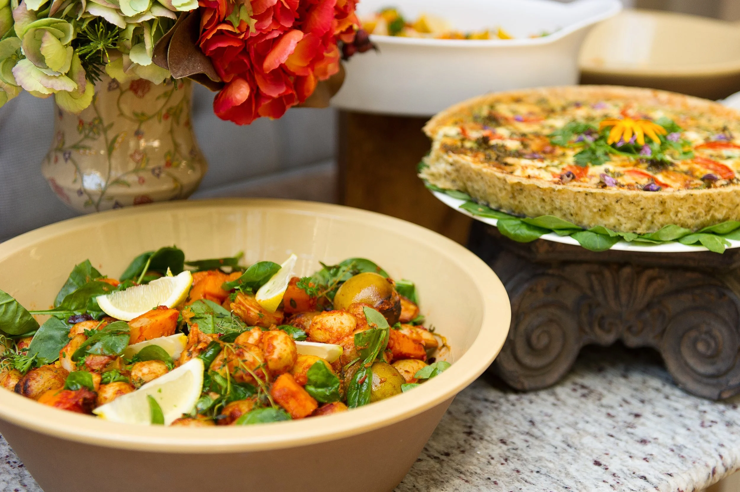 A bowl of roasted vegetables garnished with lemon wedges and fresh herbs on a stone countertop, with a floral arrangement and a layered casserole dish in the background.