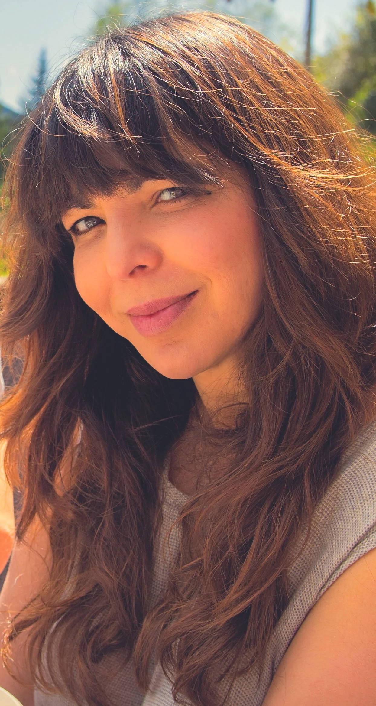 Close-up of a woman with long, wavy brown hair and bangs, smiling outdoors on a sunny day.
