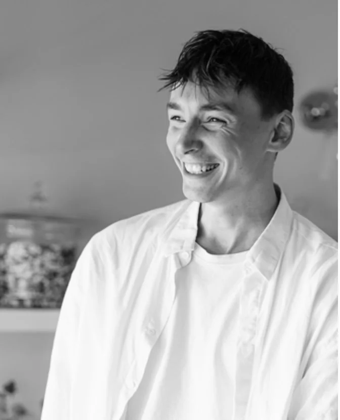 A young man with short dark hair smiling while wearing a white chef's coat in a kitchen setting.