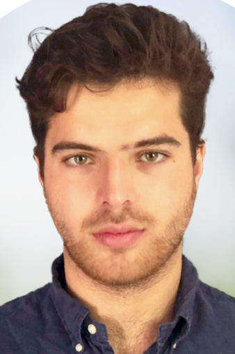 Portrait of a young man with dark hair, light skin, wearing a blue button-up shirt, against a blurred light background.