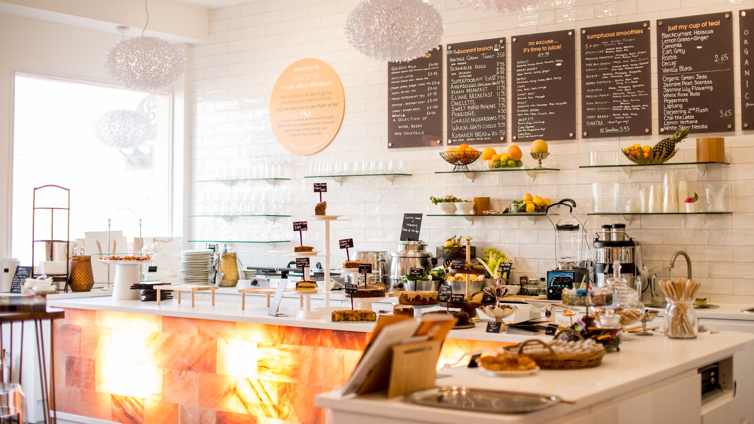 A bright, modern cafe with white brick walls and large windows. The counter displays various baked goods and desserts, with a black menu board above listing food and drink options. Decor includes fruit bowls, baskets, and sculptures, with pendant lights hanging from the ceiling.