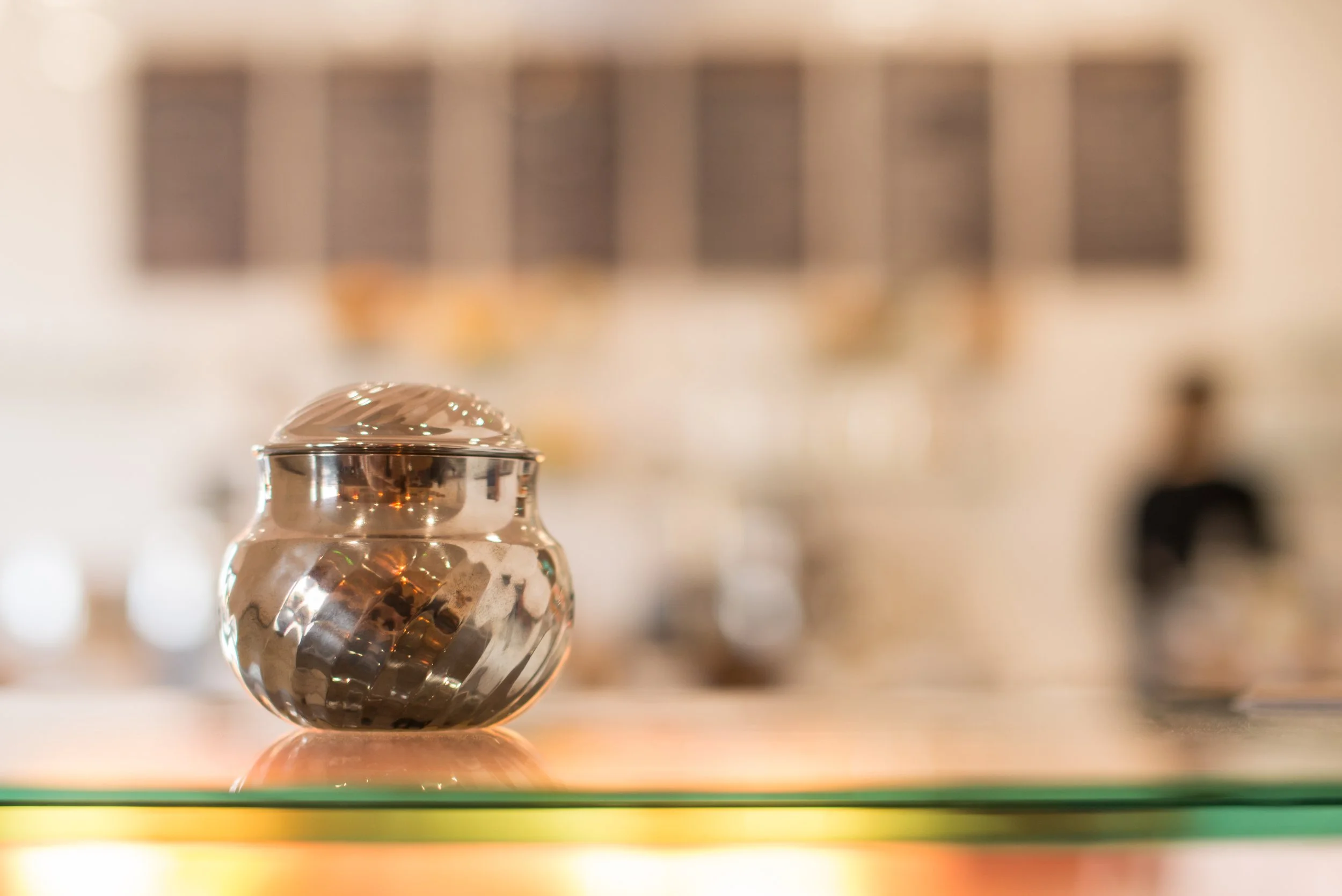 Close-up of a shiny glass marble with a reflective surface, resting on a smooth surface with colorful lighting. The background is blurred with dark and beige tones.