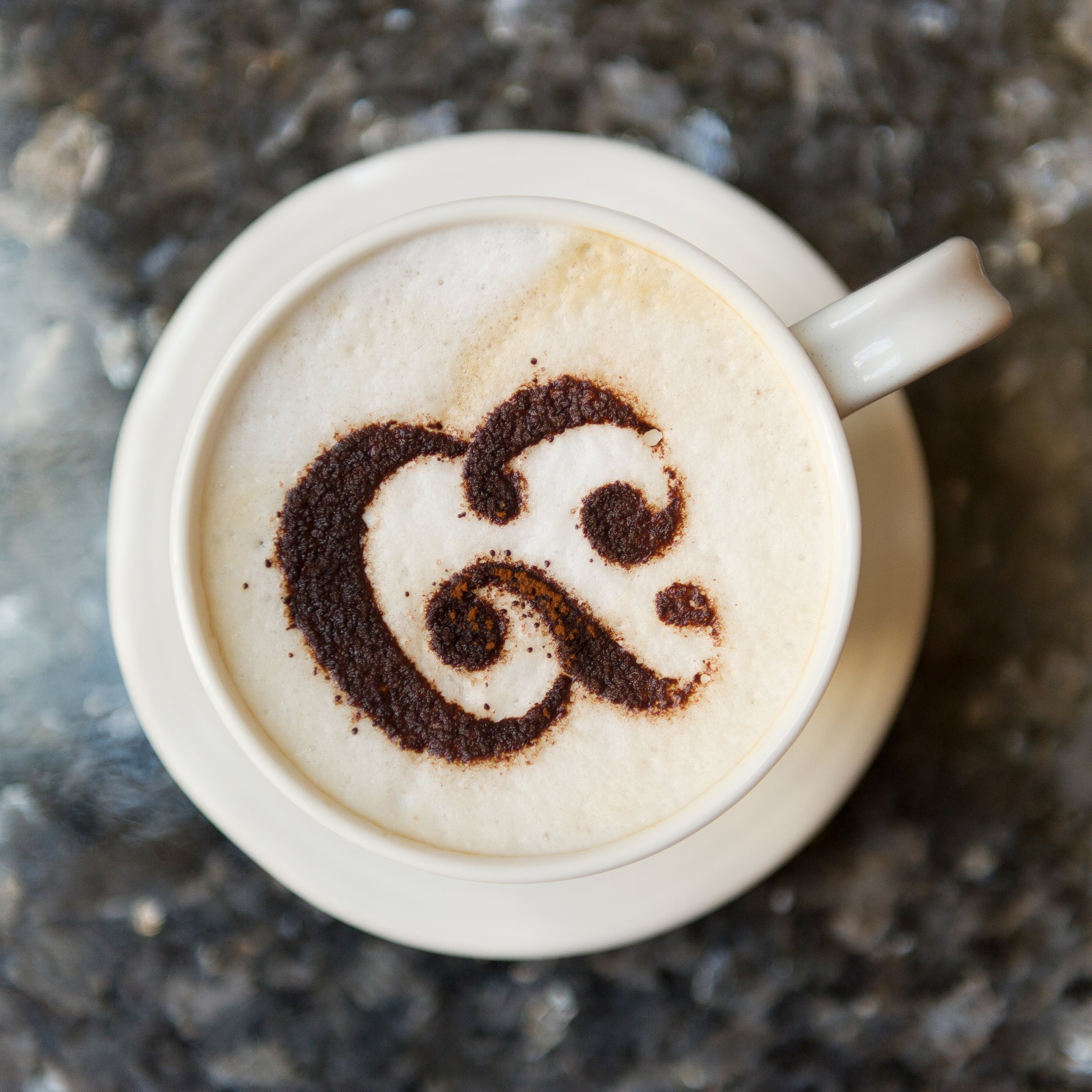 Close-up of a white mug with a dark brown coffee stain in the shape of a smiley face, sitting on a speckled black and gray surface.