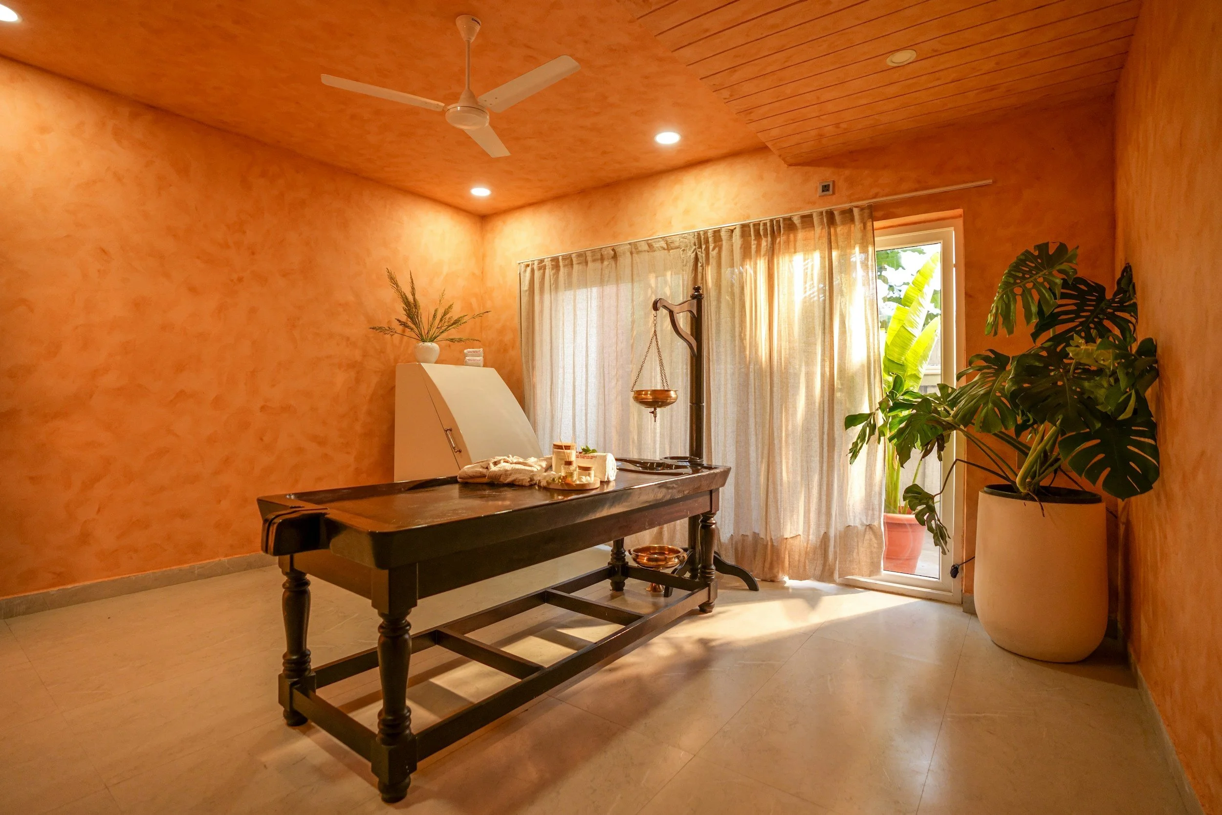 A room with orange textured walls and a ceiling with a white ceiling fan. There is a table with some items on it, a large window with beige curtains, and a large potted plant near the window.