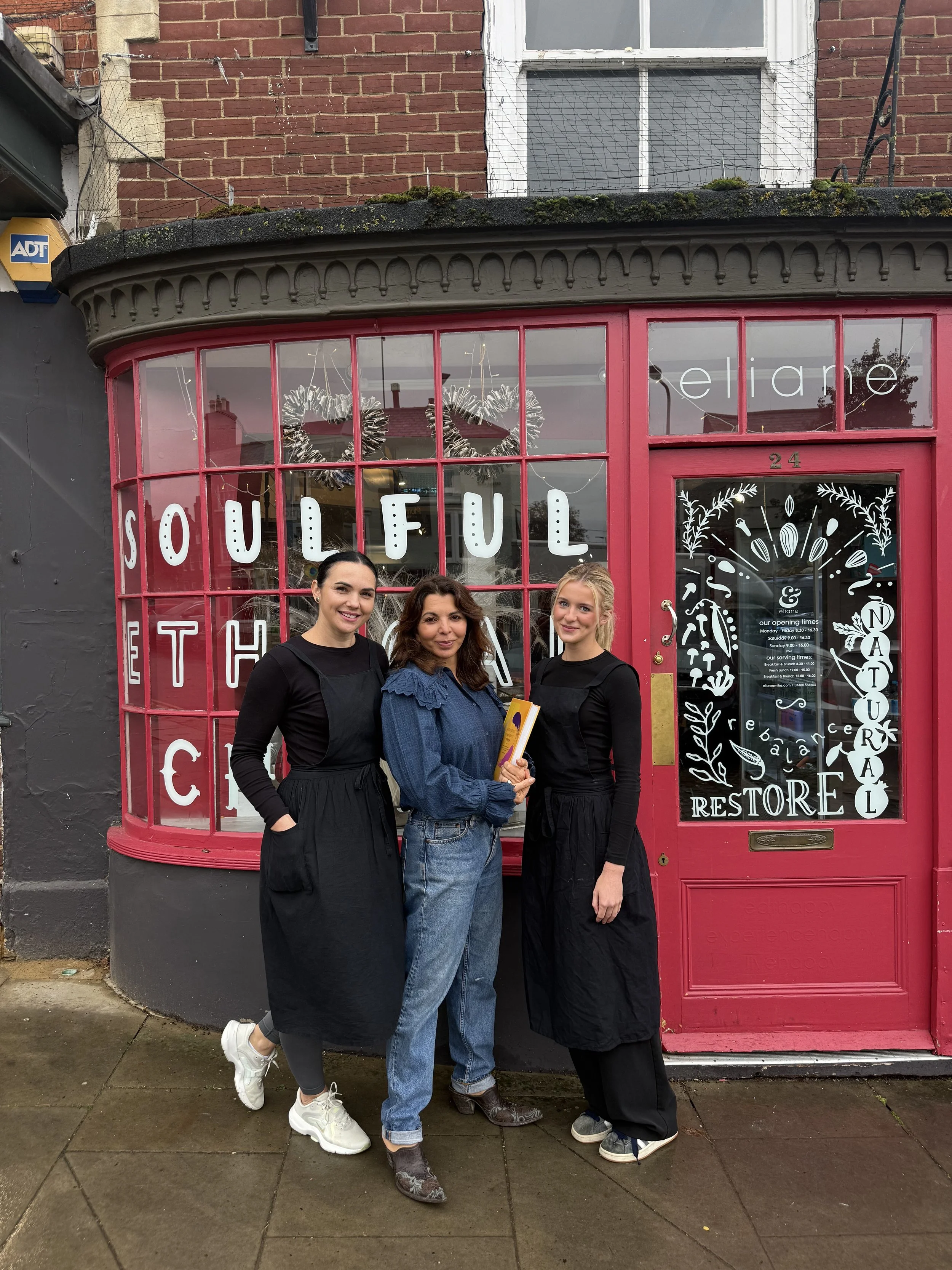 Three women standing in front of a pink storefront with large windows. The storefront has white lettering spelling out 'SOULFUL' and decorated with white illustrations of botanical elements. The women are smiling and wearing casual clothing, with two of them in black and one in blue, standing on a wet sidewalk in front of a brick building.