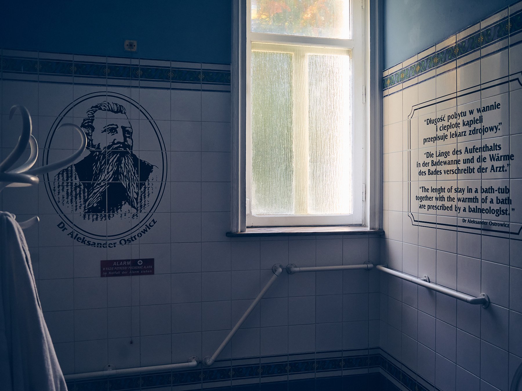 Historic tiled bathing room with a frosted window, handrails, a portrait of Dr. Aleksander Ostrowicz, and multilingual bath-treatment instructions.