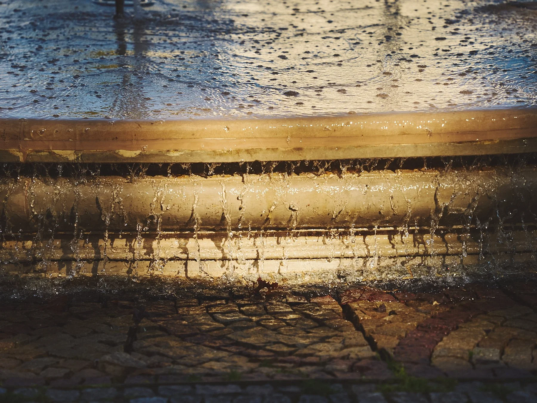 Close-up of rainwater cascading over a stone curb onto wet cobblestone pavement