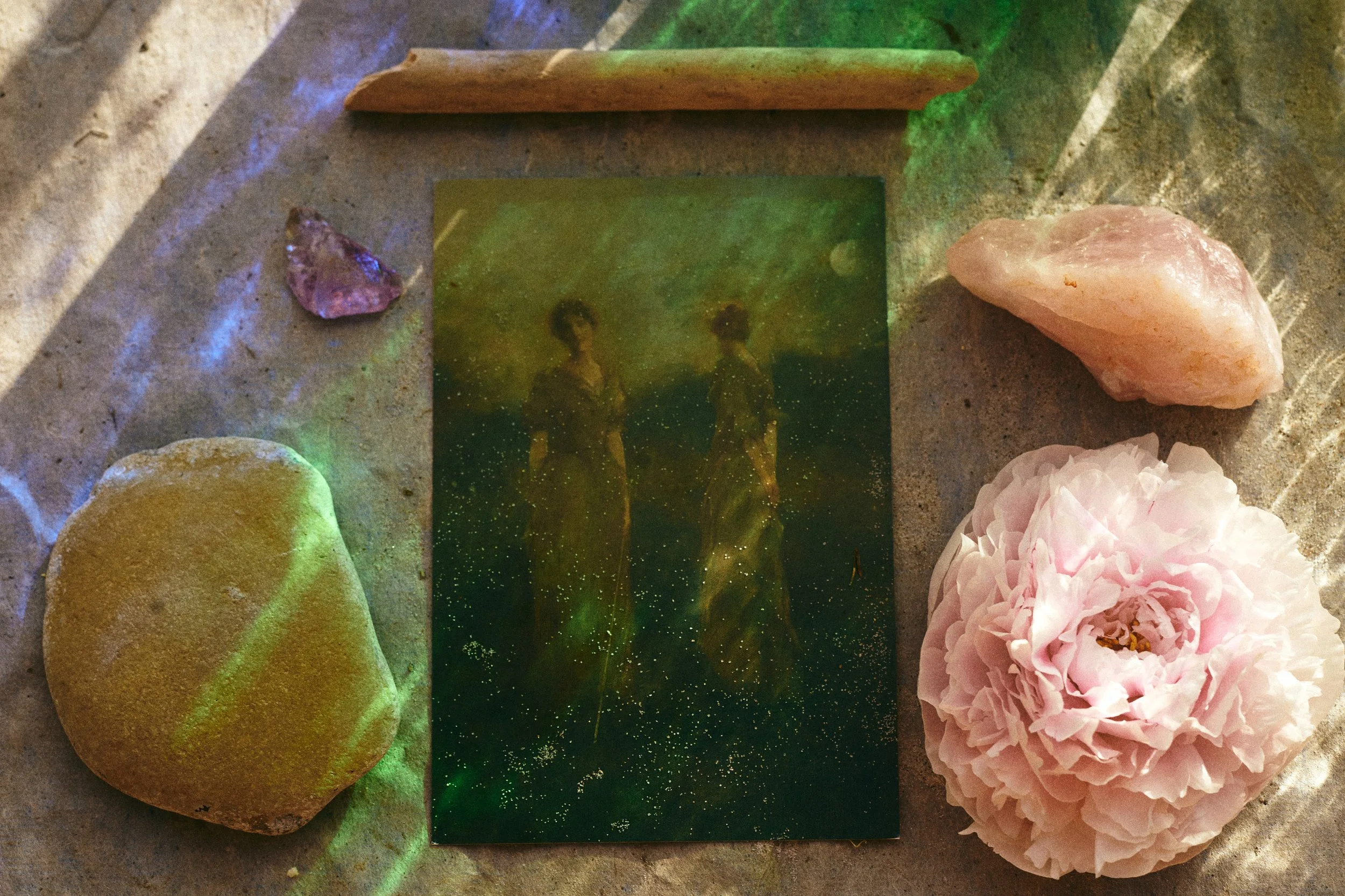 Still life of colorful stones, a dark green rectangular glass tile, wooden pieces, and pink flowers on a textured surface