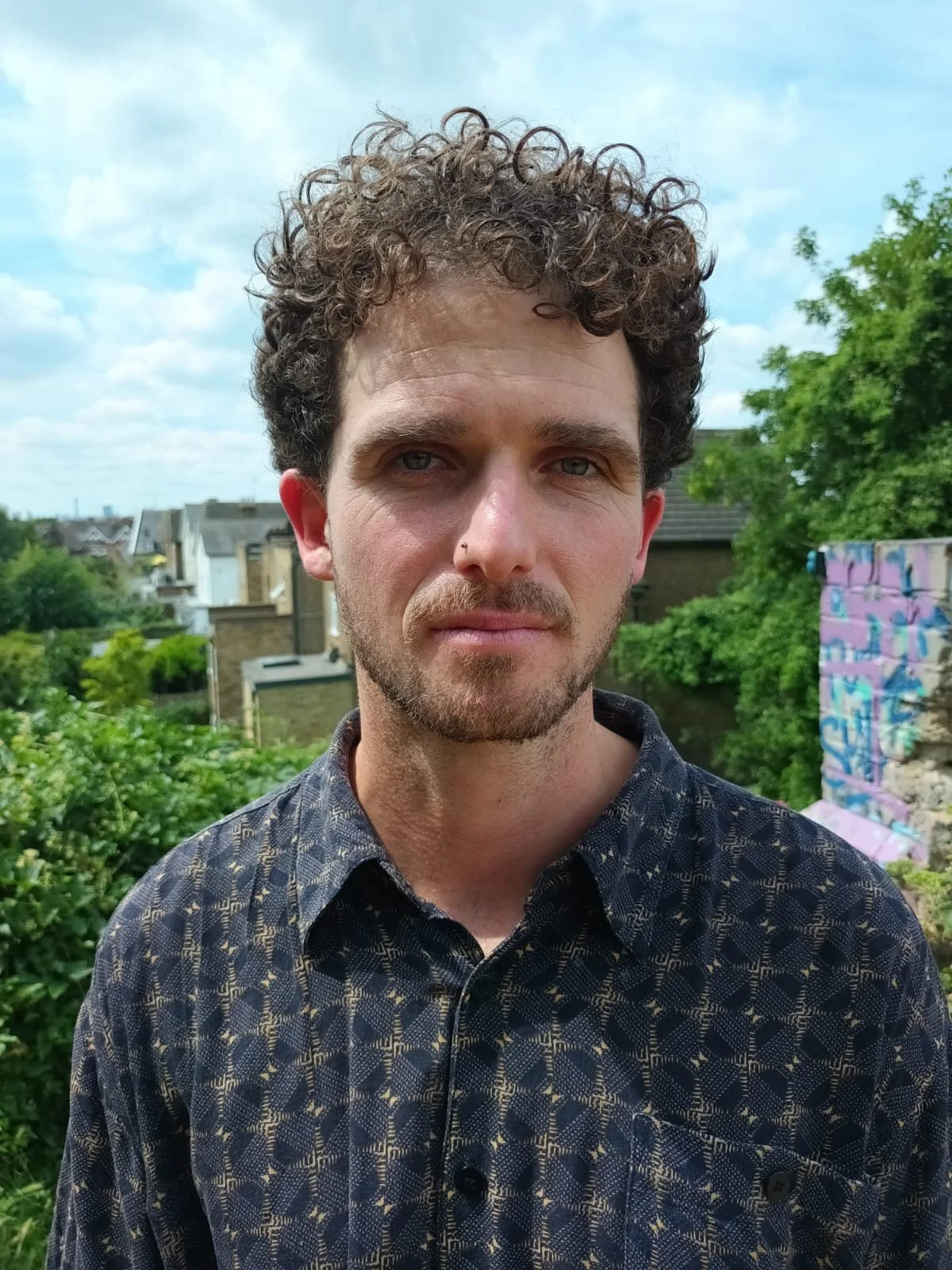 A man with curly brown hair and a beard stands outdoors, wearing a patterned button-up shirt. There are trees, buildings, and a graffiti-covered wall in the background under a partly cloudy sky.
