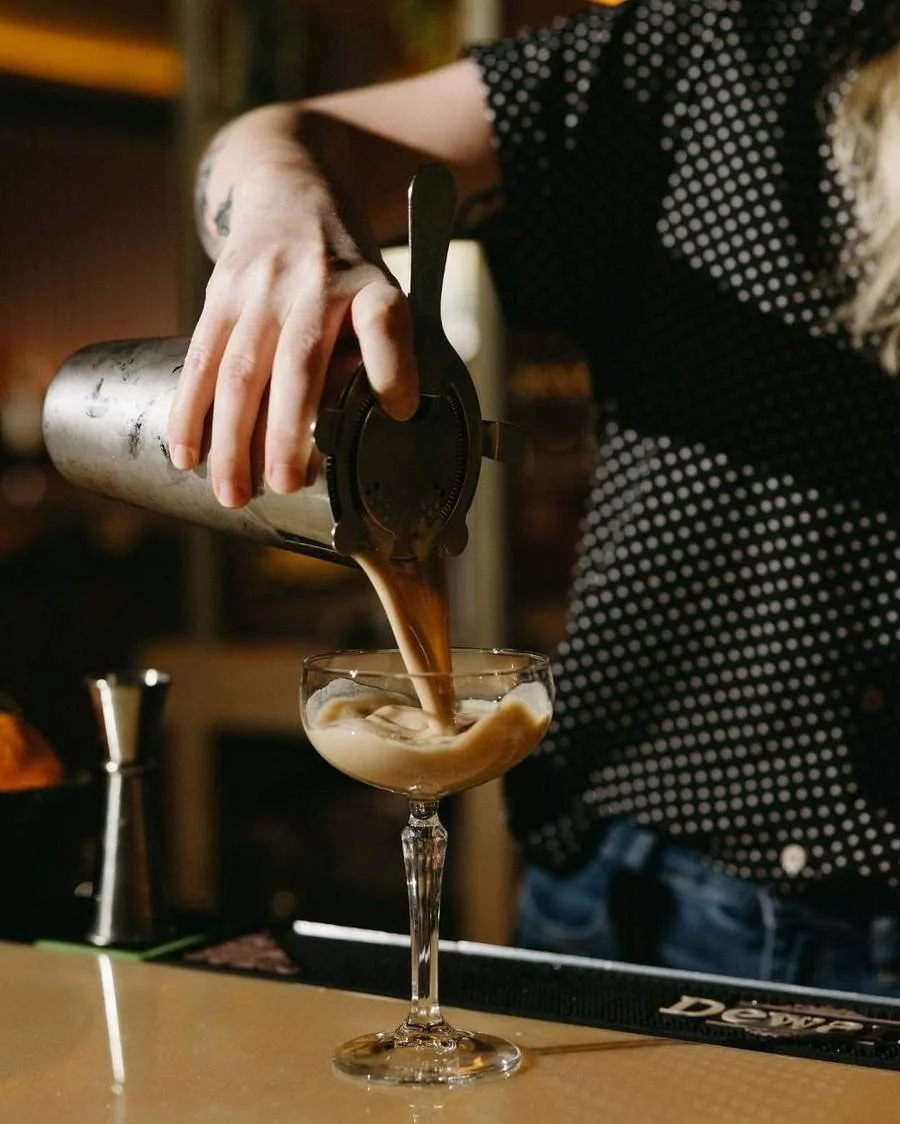 Bartender pouring a creamy cocktail from a metal shaker into a coupe glass
