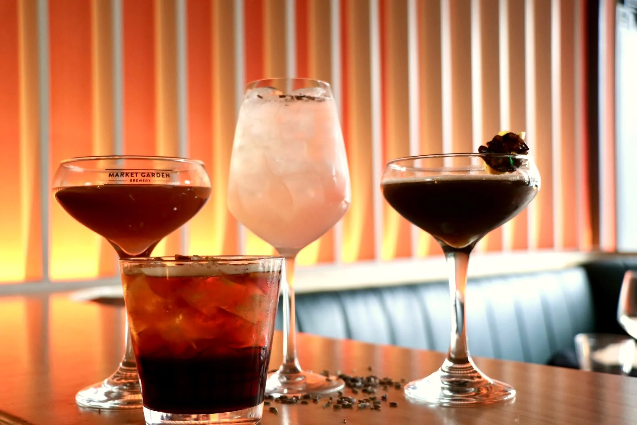 Four assorted cocktails and beer-style drinks in glasses on a table, including dark drinks, a frothy pale drink, ice, and garnishes.