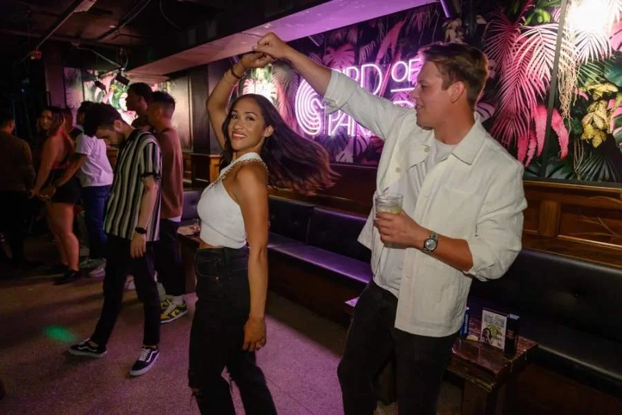 Couple dancing under neon lights at a tropical-themed nightclub while other people socialize nearby.
