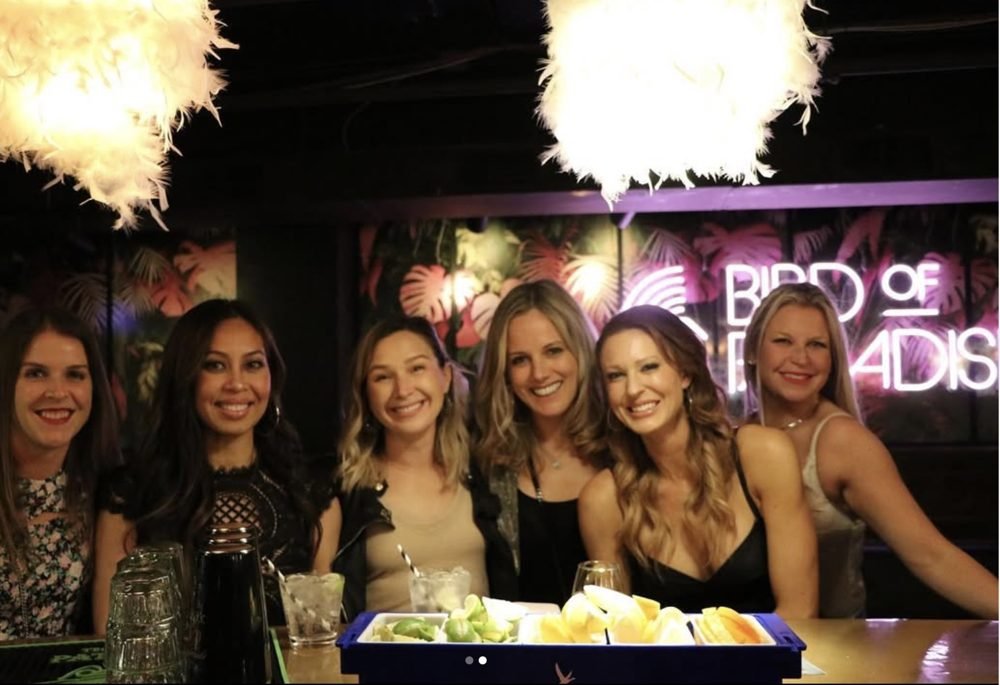 Group of five women posing at a restaurant bar with drinks and a neon “Bird of Paradise” sign
