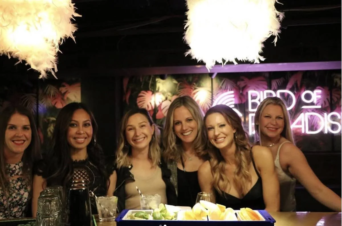 Six women posing together at a restaurant table beneath a neon “Bird of Paradise” sign.