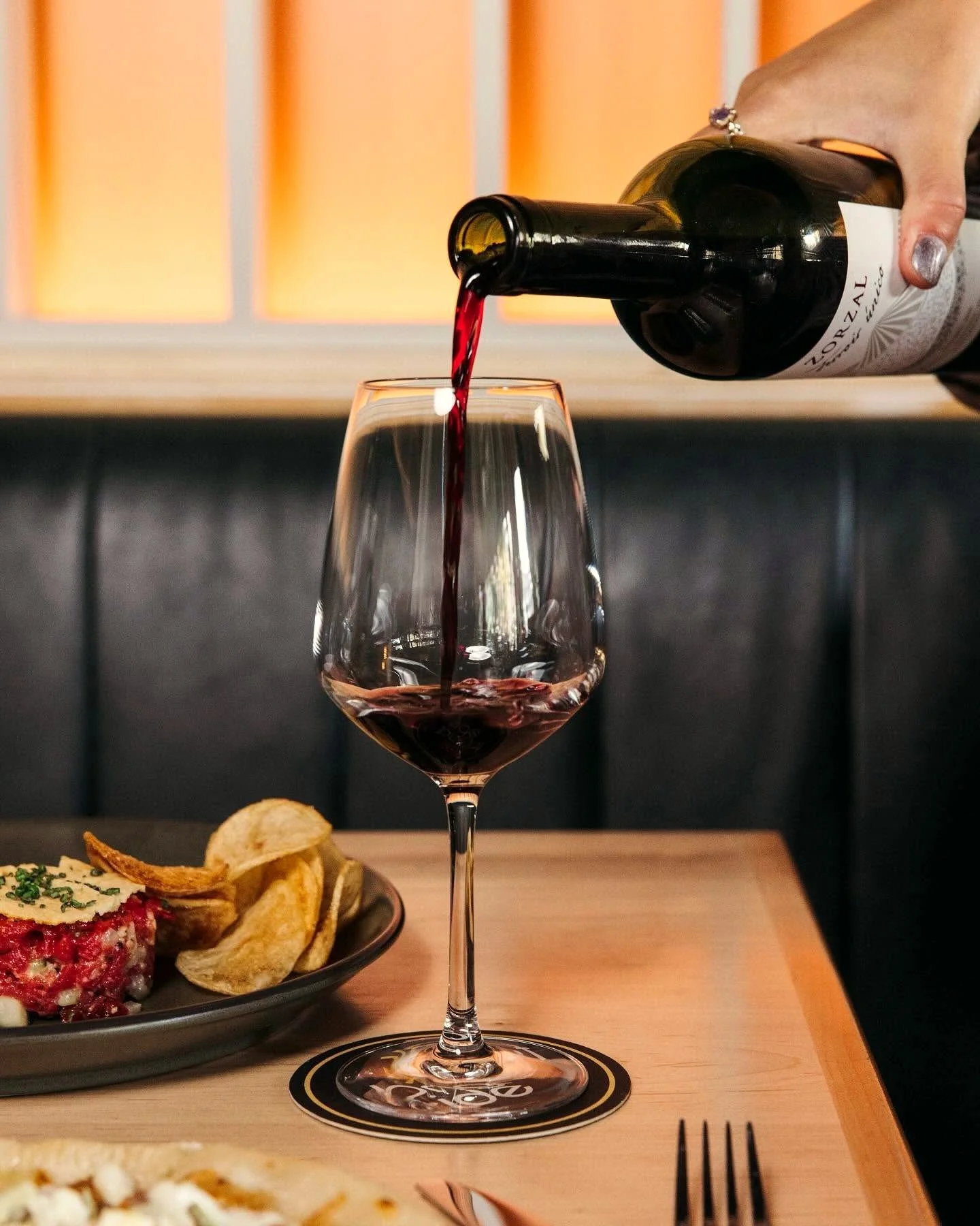 Red wine being poured from a bottle into a wine glass beside a plate of chips and appetizer