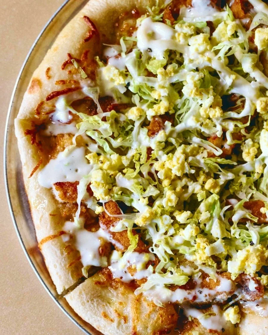 Close-up of a loaded pizza topped with lettuce, scrambled egg, crispy bacon, melted cheese, and white sauce.
