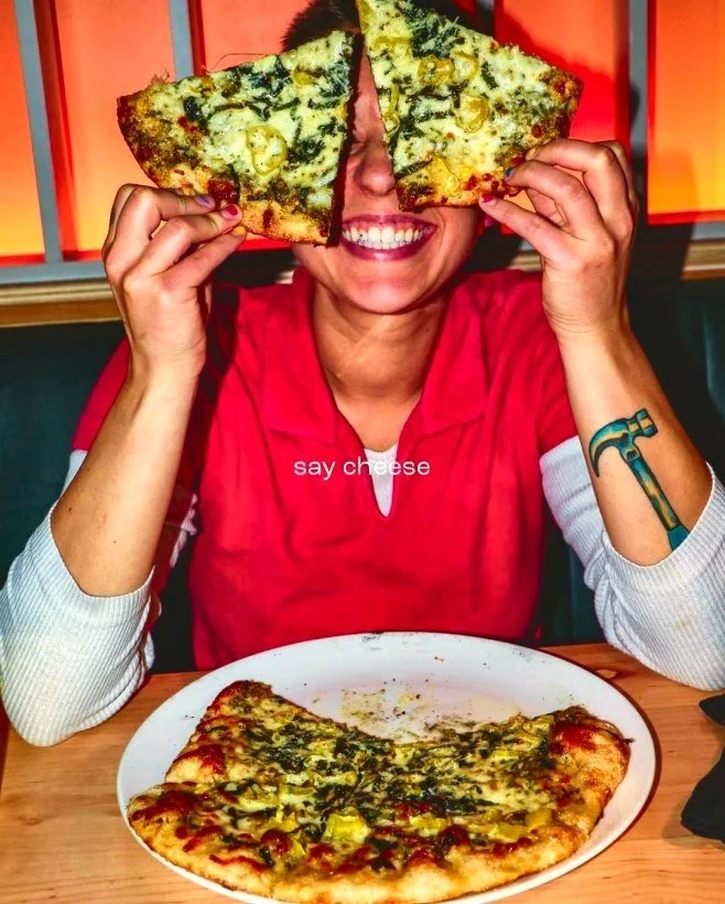 Smiling woman in a red shirt holds two slices of vegetable pizza over her eyes at a restaurant table.