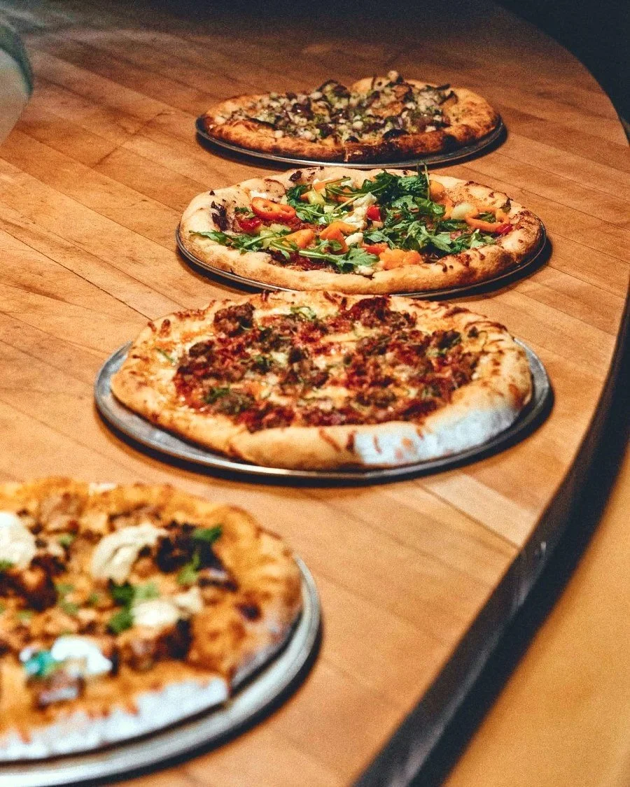 Four assorted artisan pizzas on metal trays atop a wooden table, including vegetable-topped and cheese pizzas.