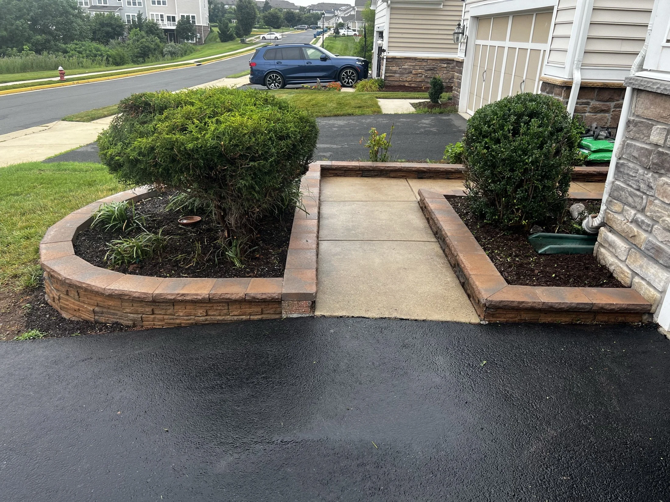 Landscaped townhouse entry with brick retaining walls, concrete walkway, trimmed shrubs, driveway, and parked blue SUV