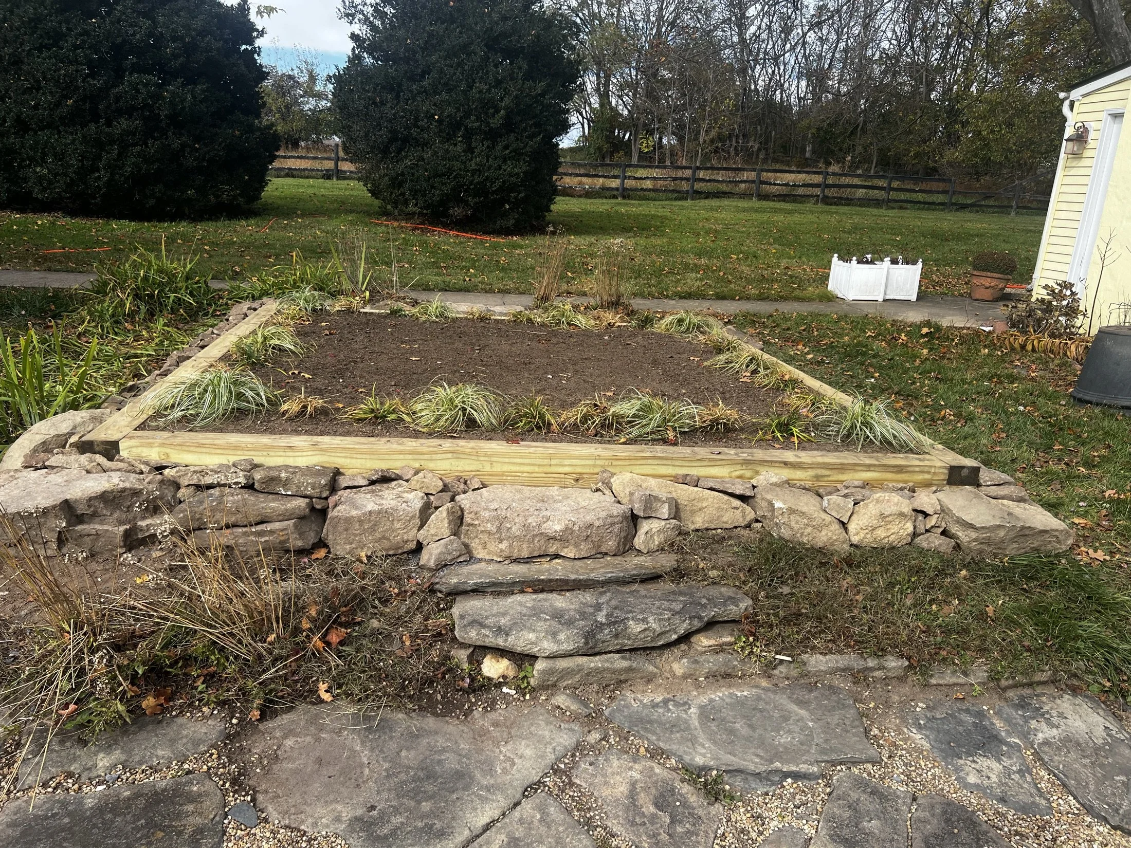 Landscaped backyard with a raised stone-and-wood garden bed, ornamental grasses, lawn, trees, and a wooden fence.