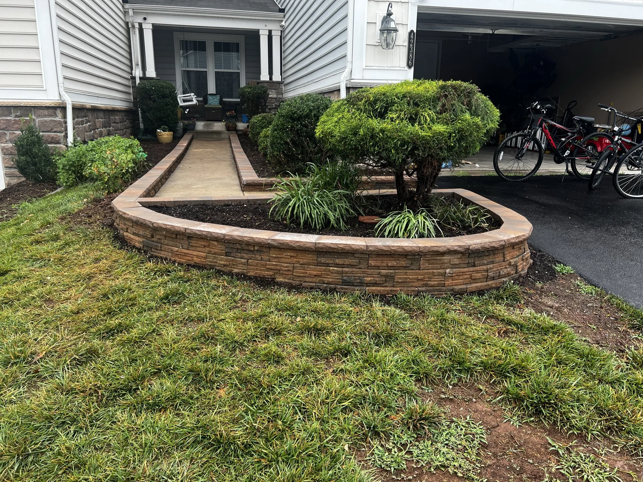 Landscaped front yard with a stone retaining wall, shrubs, walkway, house entrance, and bicycles in an open garage