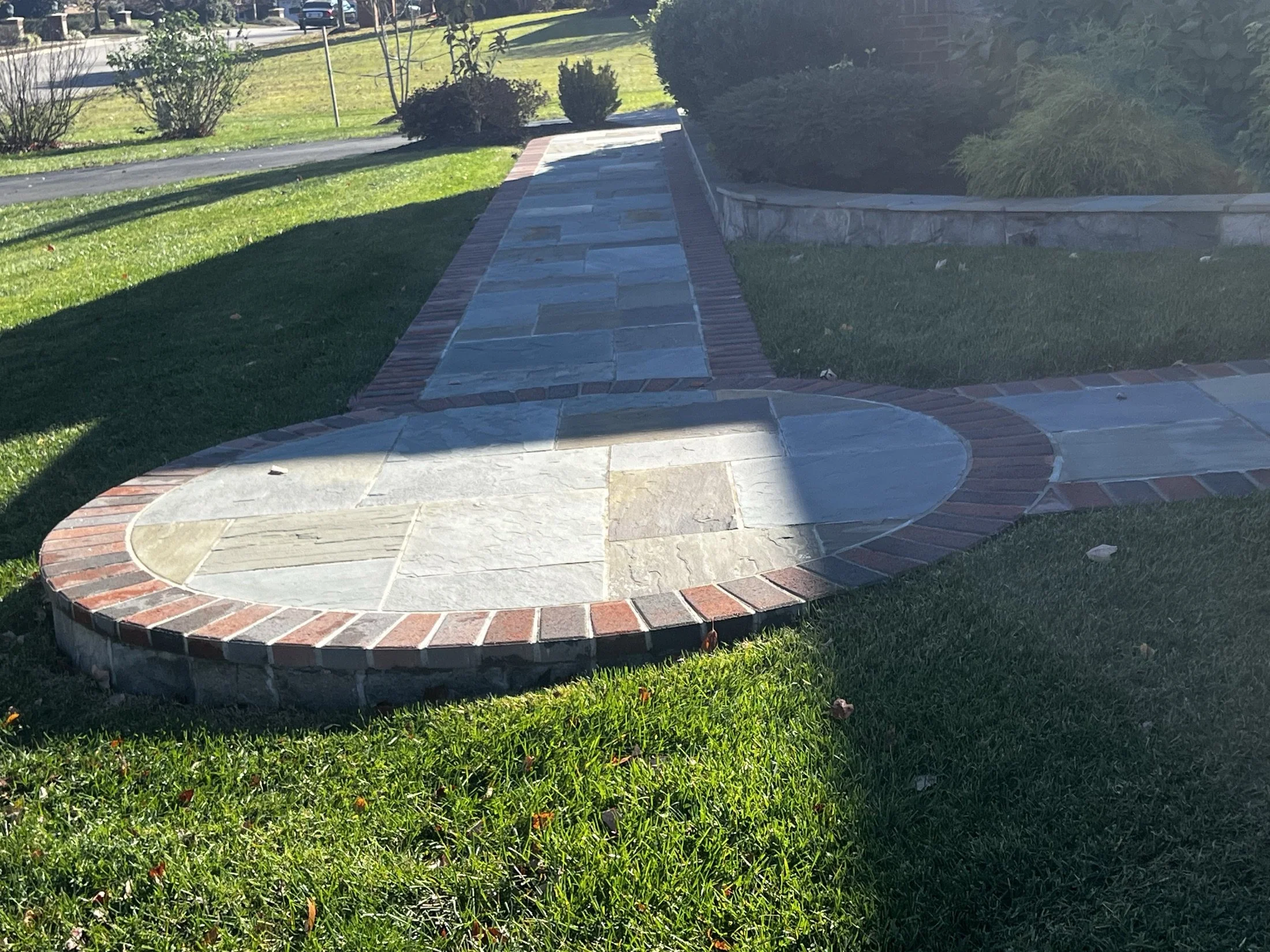 Landscaped front yard with a circular flagstone patio, brick edging, walkway, lawn, shrubs, and stone retaining wall.