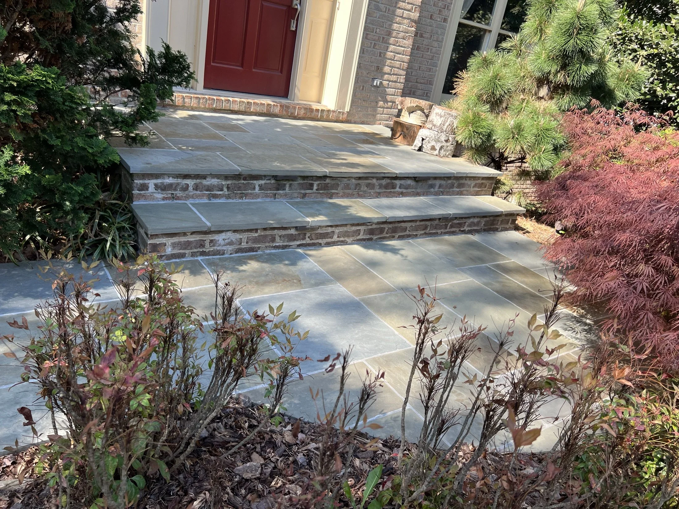 Brick home entry with a red door, slate stone steps, patio, and landscaped shrubs