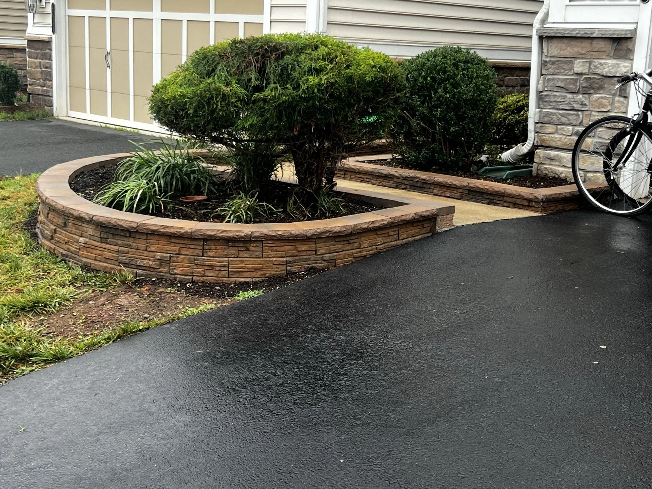 Landscaped residential driveway with stone-edged garden beds, trimmed shrubs, ornamental grasses, garage, and a bicycle.