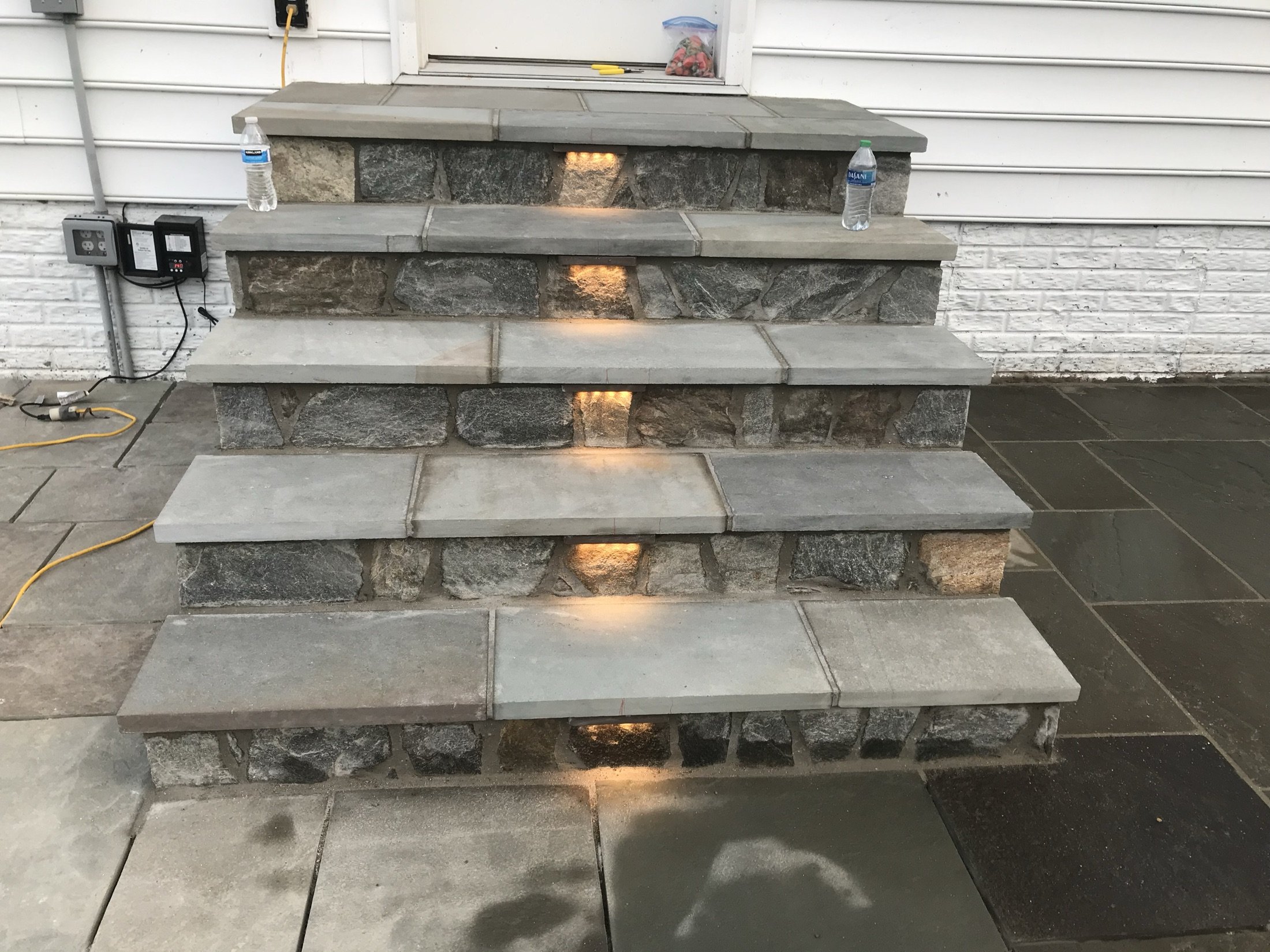 Tiered stacked-stone and concrete outdoor fireplace with glowing recessed lights, water bottles, and electrical controls