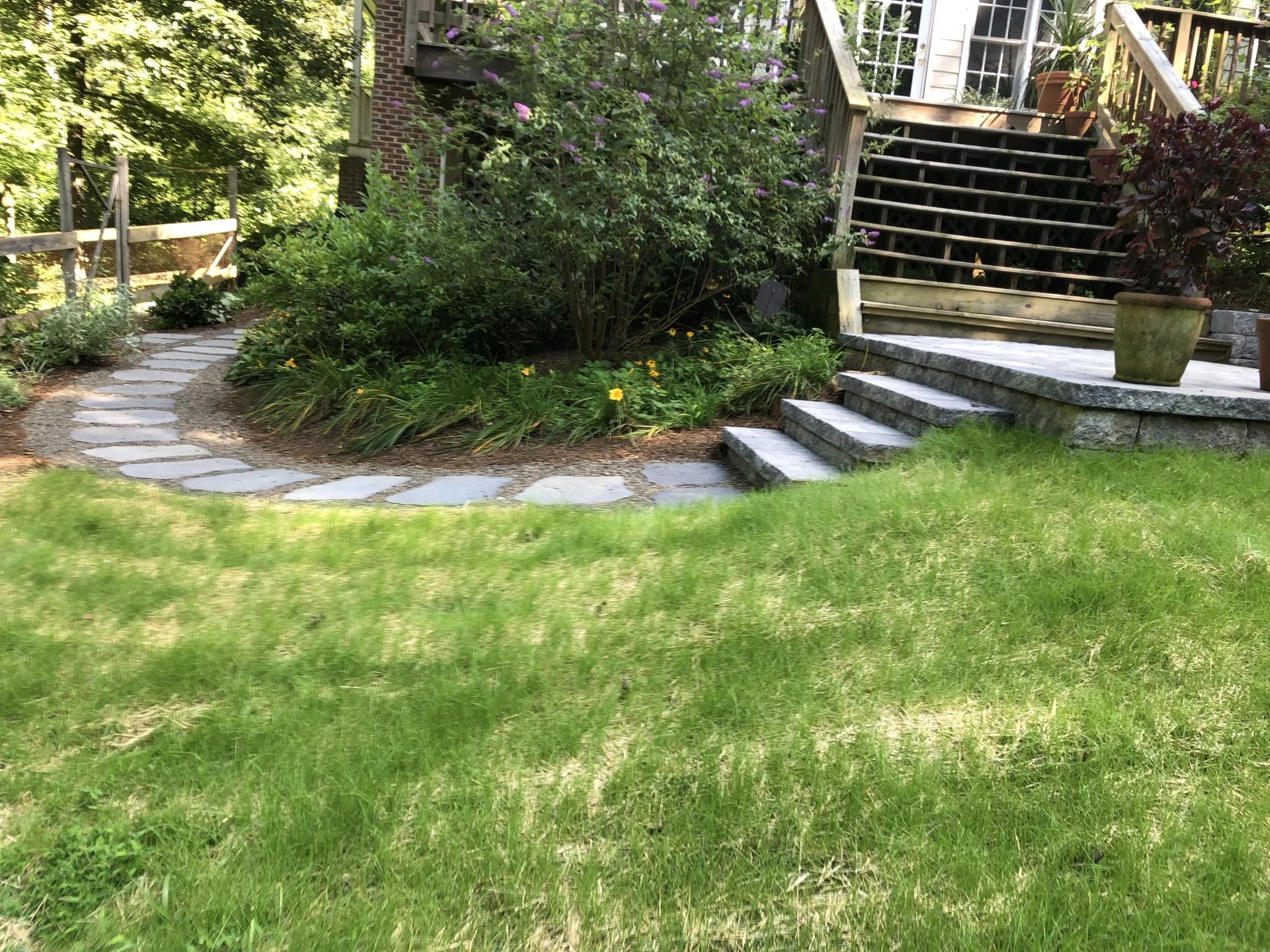 Landscaped backyard garden with stepping-stone path, flowering shrubs, stone steps, and wooden deck stairs