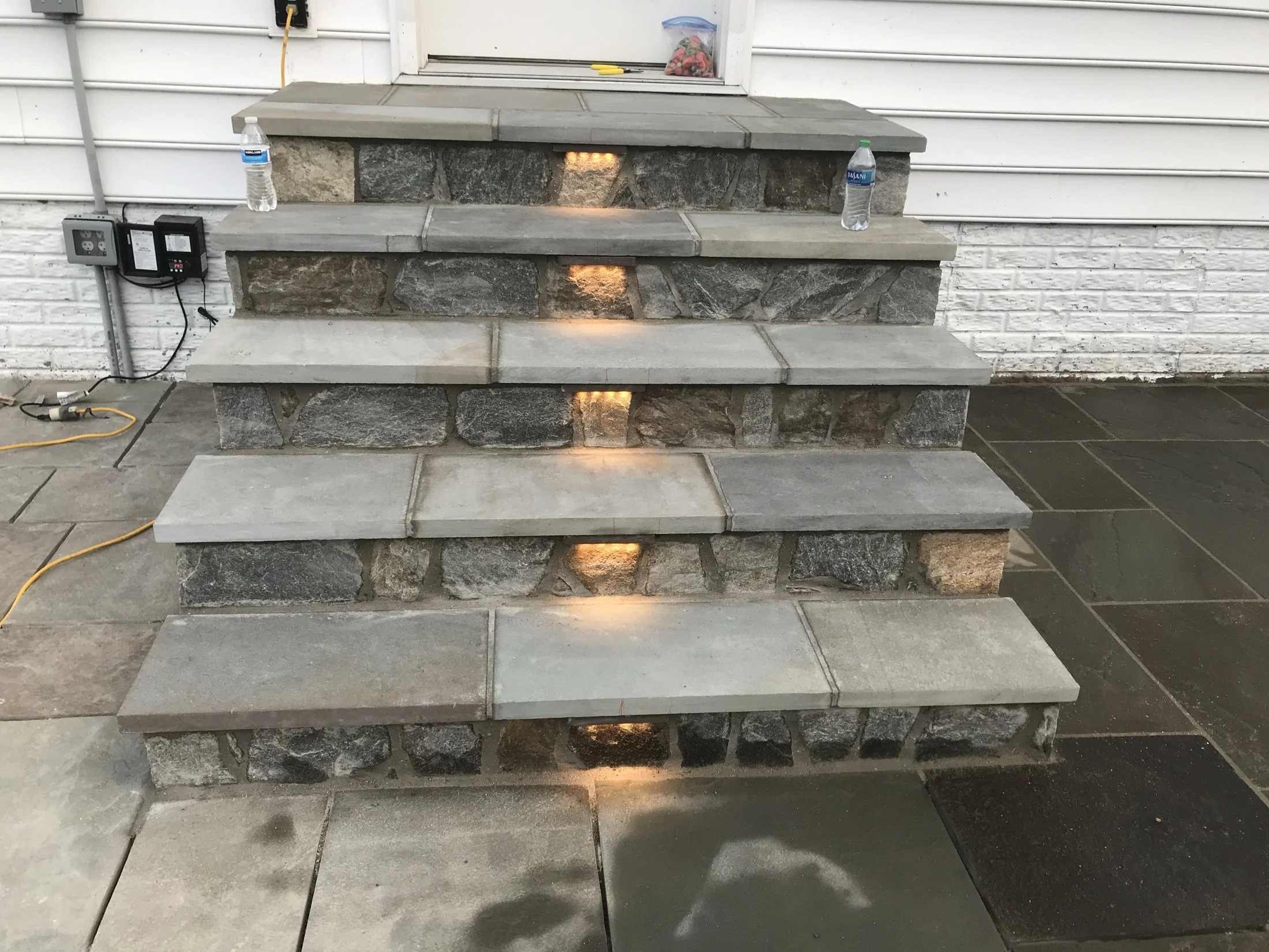 Tiered stone fireplace with glowing recessed lights, two water bottles, and electrical cords in a tiled outdoor area