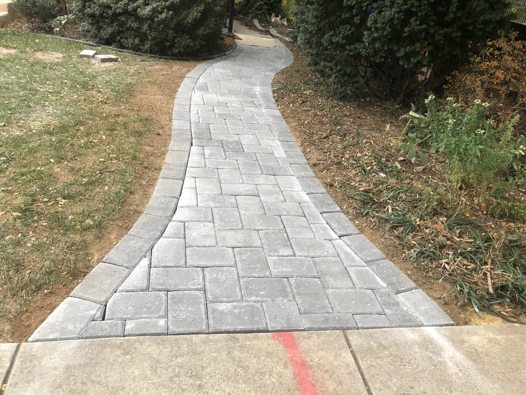 Curved gray paver walkway winding through a landscaped garden with grass, shrubs, and trees